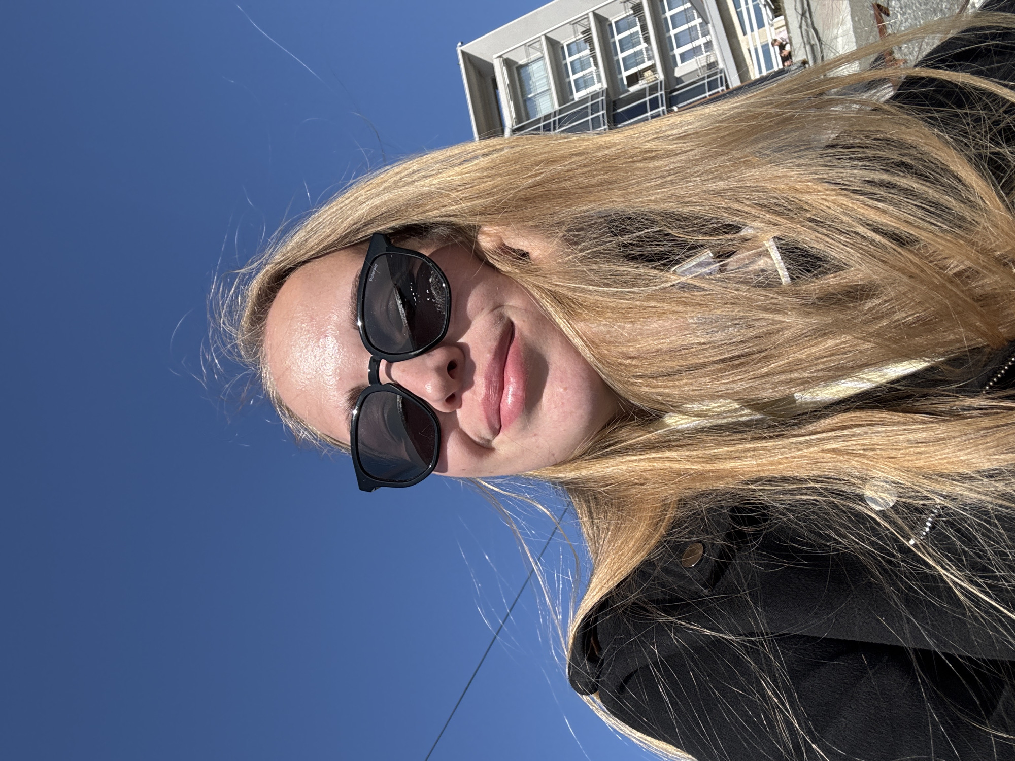 A close-up, slightly tilted portrait of a young woman is captured outdoors in Sochi, Russia, under a clear blue sky on a bright, sunny day. The woman has long, straight blonde hair that extends across much of the frame. She wears black sunglasses, with the brand "Polaroid" visible on the left temple, obscuring her eyes. Her lips are slightly parted, suggesting a subtle smile or pout. She is dressed in a black jacket or coat, visible on her right side, featuring some metallic studs or buttons. The background is dominated by a vibrant blue sky. In the upper right portion of the image, part of a modern building with a contemporary architectural style, characterized by multiple windows and balconies, is visible. Faint power lines or cables traverse the sky horizontally in the mid-left area. The scene conveys a casual, bright, daytime atmosphere.