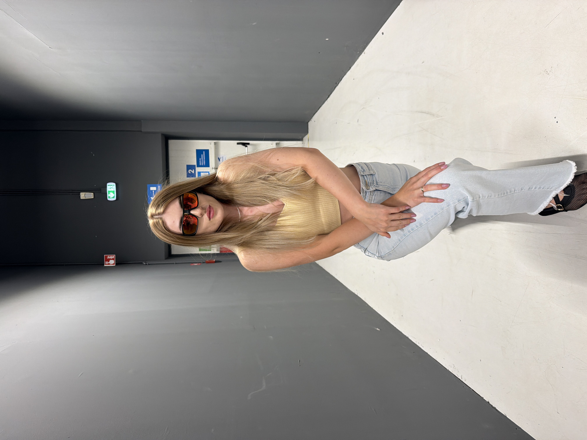 A young woman with long blonde hair and sunglasses is kneeling on a white floor in a narrow hallway with grey walls. She is wearing a yellow crop top and light blue jeans. There are signs on the walls, including a red fire extinguisher sign and a green exit sign. The hallway appears to be indoors and the lighting is artificial.