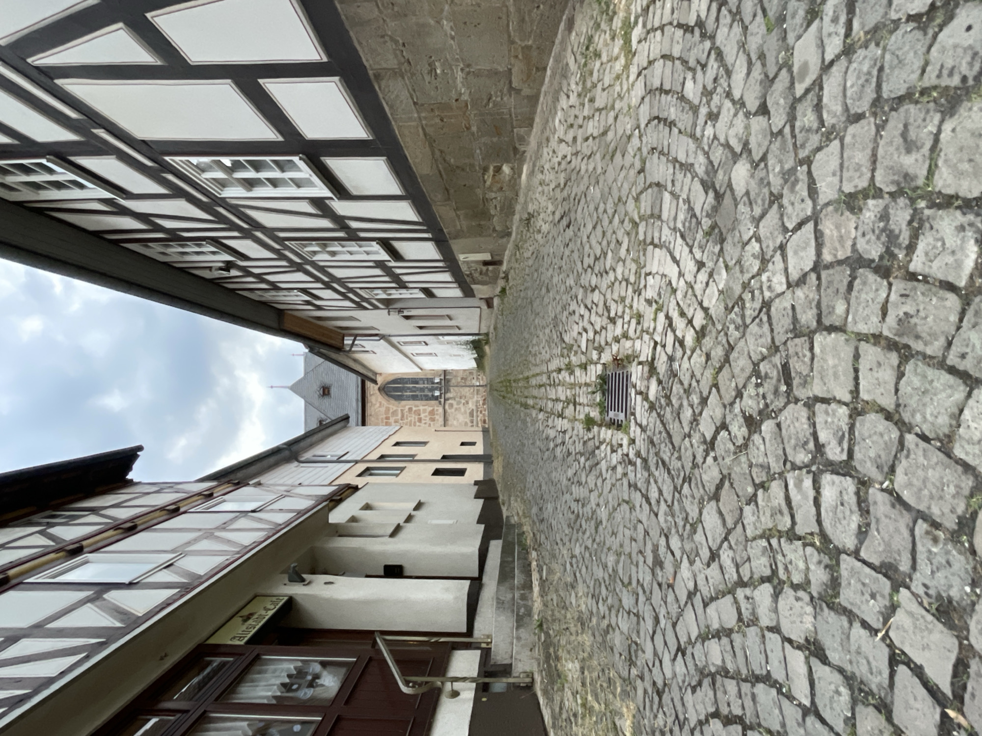 The image depicts a cobblestone street in the historic town of Neukirchen, Germany. The street slopes upwards, leading towards a building with a pointed steeple, possibly a church, in the distance. Flanking the street are traditional timber-framed buildings, characteristic of German architecture. The building on the left has a sign that reads "Altstadt-Cafe," indicating a cafe in the old town. The sky is overcast, suggesting a cloudy day. The scene is devoid of people or any overt activity, presenting a quiet, almost static view of the street. The focus is on the architecture and the texture of the cobblestones.