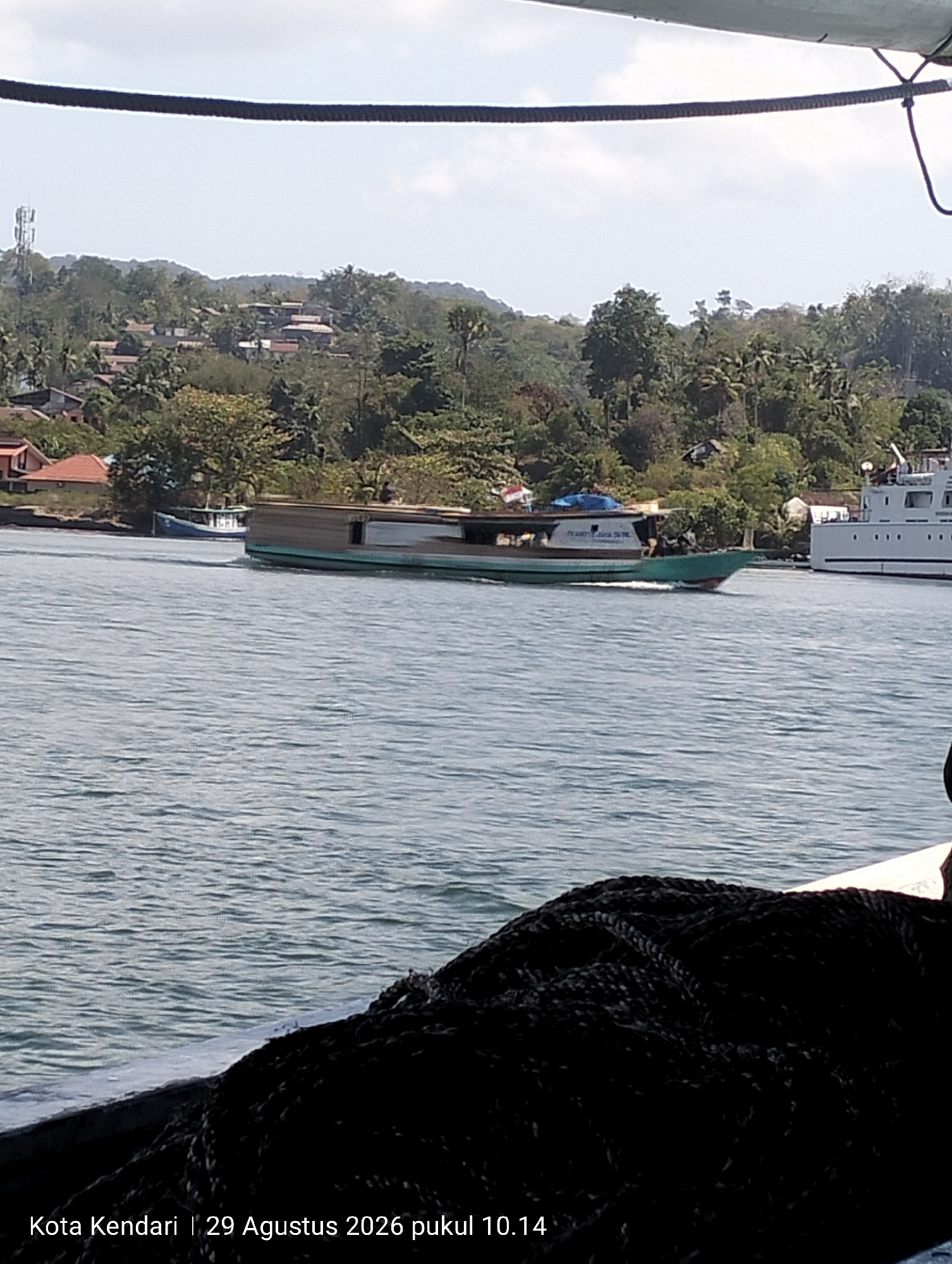 In Kendari, Indonesia, on August 29, 2026, at 10:14 AM, a wide-angle shot captures a body of water with a partially visible boat in the foreground, laden with dark ropes. In the mid-ground, a green and brown passenger boat is navigating the water, leaving a wake. To the left of the passenger boat, a smaller blue boat is visible near the shoreline. The background features a lush, green hillside dotted with houses and vegetation. A cell tower is situated atop the hill. Further to the right, a white ferry or ship is docked. The sky is bright and partly cloudy.