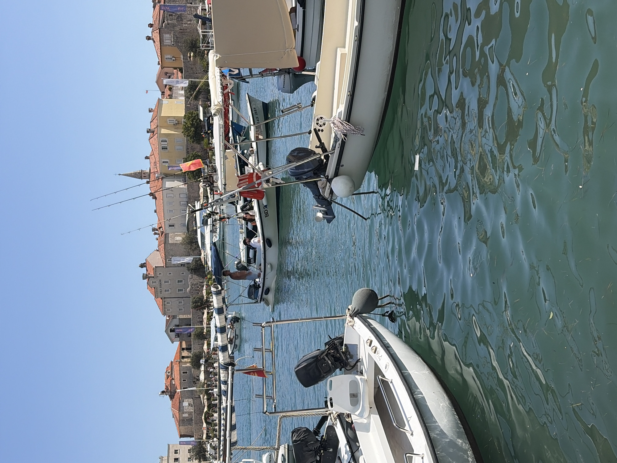 The image shows a harbor in Budva, Montenegro, with several boats docked in the clear blue water. In the background, a historic town with red-tiled roofs and stone buildings lines the waterfront under a bright, cloudless sky. The foreground is dominated by the side of a white boat, its metallic railing visible, and a dark outboard motor at the stern. Another boat, also white with a cream-colored canopy, is moored to its right. Several other boats, some with people on board, are scattered throughout the harbor. Fishing rods are visible on at least two boats, suggesting recreational fishing activities. The overall scene depicts a sunny day in a picturesque coastal town.
