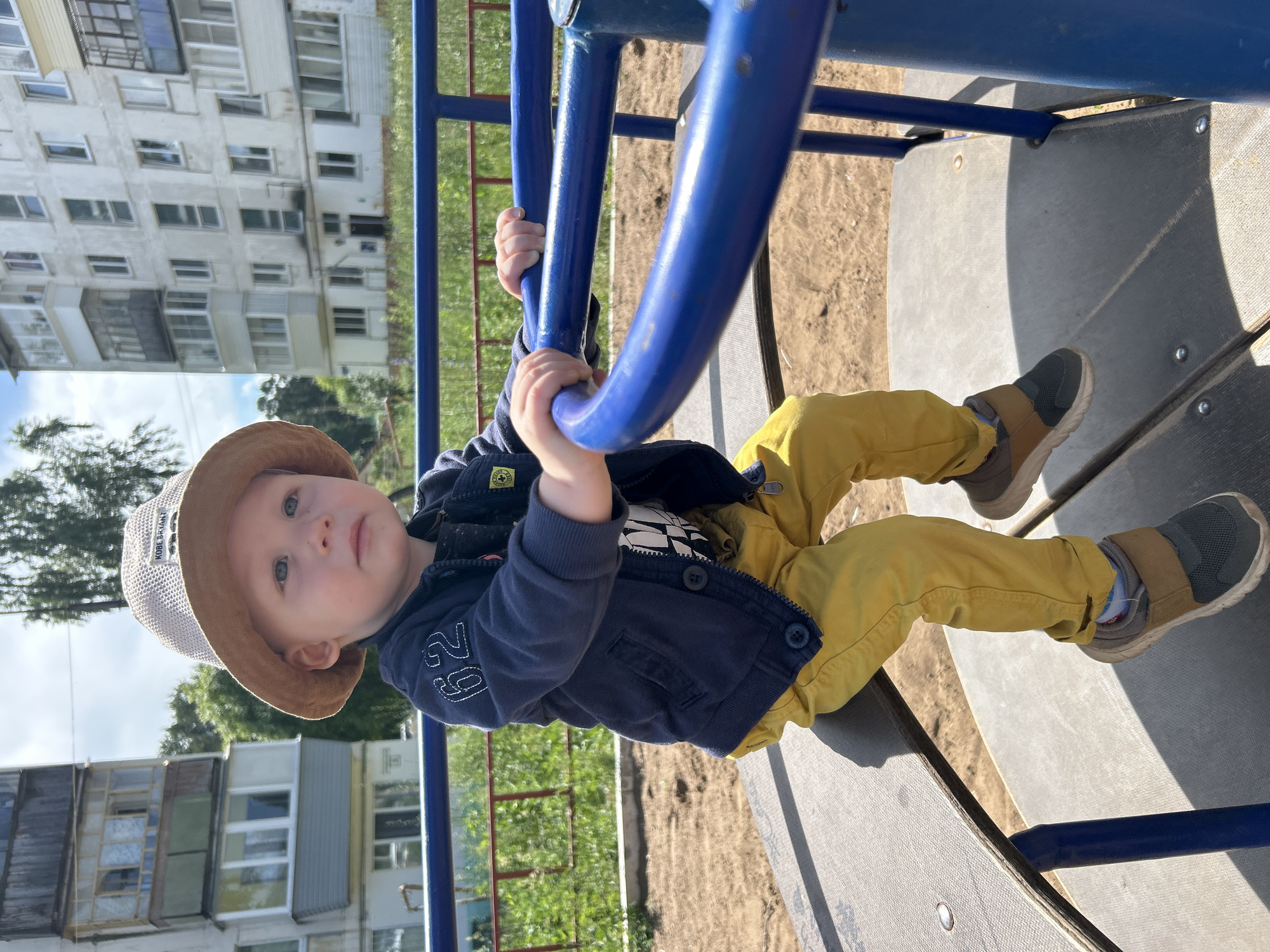 A young boy wearing a beige bucket hat, a dark blue jacket with white trim number "62" on the sleeve, and yellow pants sits on a merry-go-round at a playground. He is holding onto the blue metal bars of the ride. The merry-go-round is in a sandy area with green grass and trees in the background. Behind the playground, a light-colored apartment building is visible. The overall scene suggests a sunny day at a residential playground in Chaikovsky, Russia.