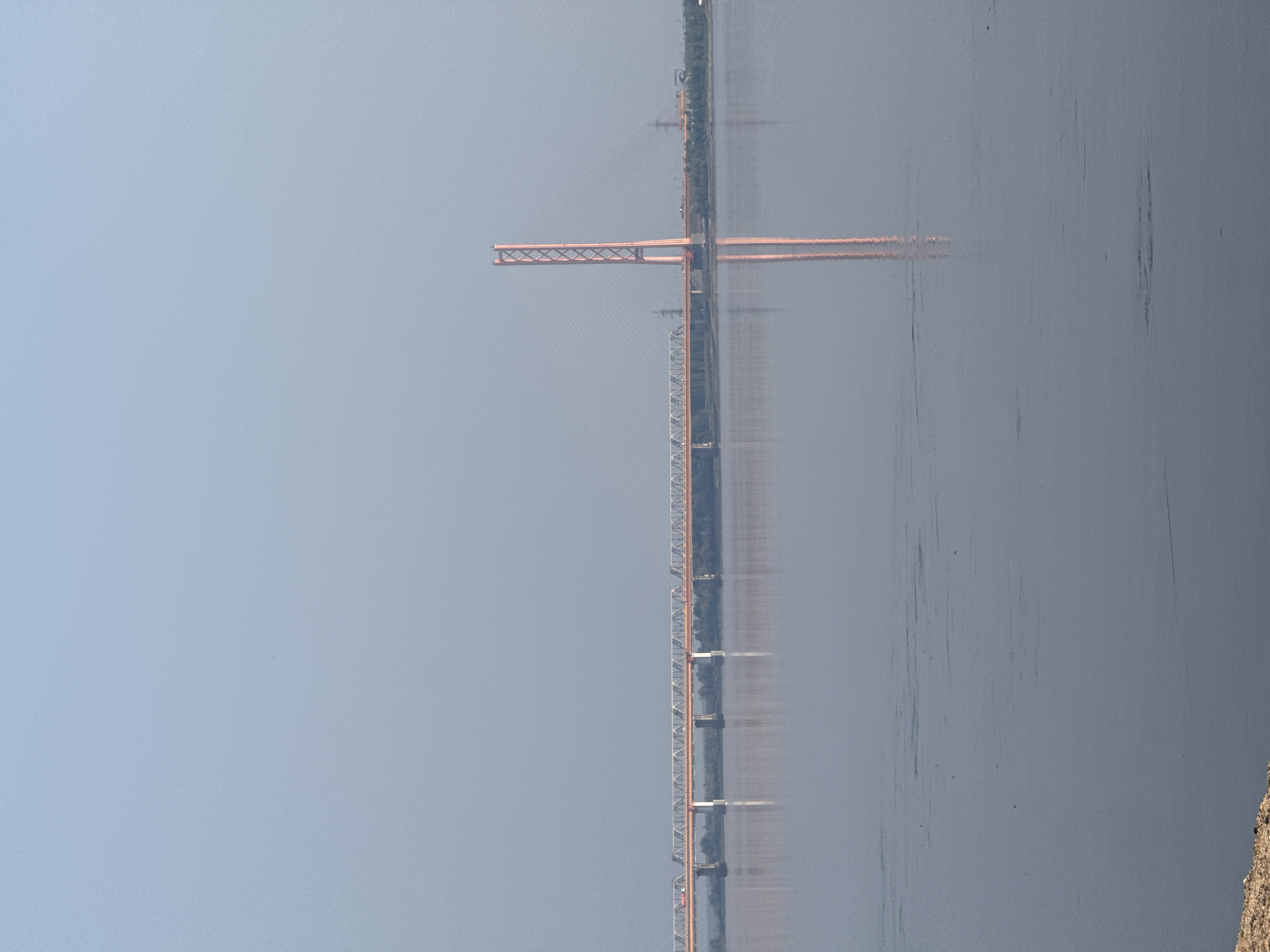 The image shows a wide, calm body of water, likely a river, reflecting a hazy, pale blue sky. A long, multi-span bridge stretches horizontally across the middle ground. This bridge appears to be a truss bridge with a light-colored deck supported by white pylons. On the right side of the frame, a single, distinctive cable-stayed bridge tower, colored in a muted reddish-orange hue with a lattice-like structure, rises prominently. Its reflection is clearly visible in the water below. The far bank of the river is lined with a dense strip of trees, with some industrial structures and power lines visible behind them. In the foreground, the edge of a sandy or rocky embankment is visible on the bottom left. The overall atmosphere is serene and somewhat muted due to the soft lighting and atmospheric haze. This scene depicts a bridge crossing over the Irtysh River near the city of Barsovo, Russia, a region known for its industrial and port activities.