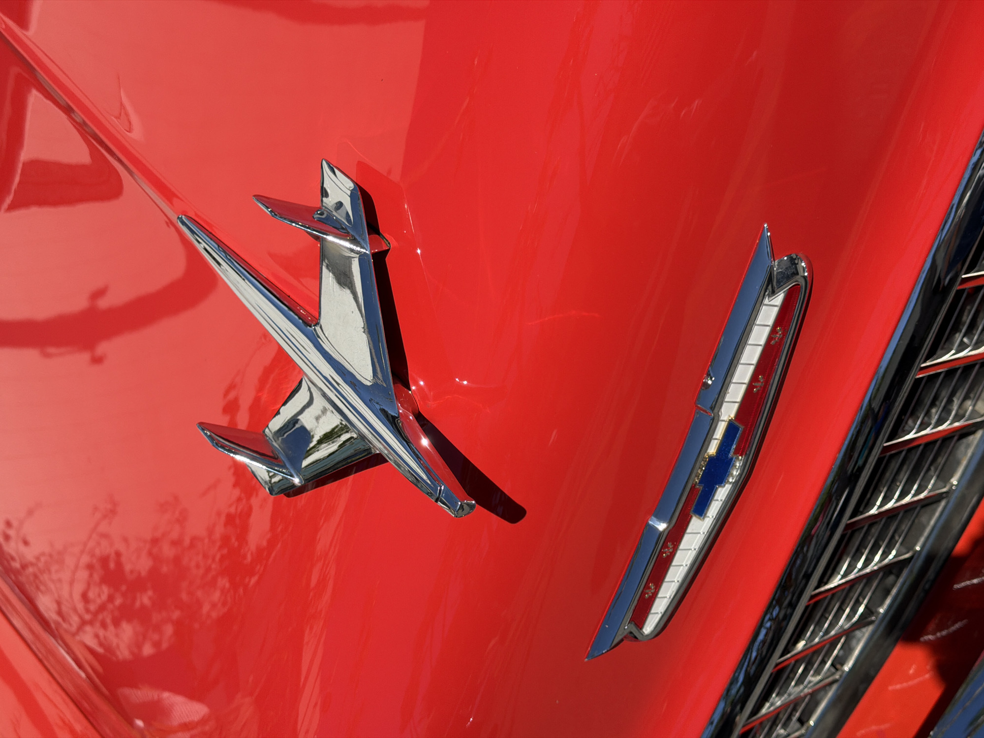 The image presents a detailed, close-up view of the front hood of a vintage red automobile. The highly polished, vibrant red paint dominates the frame, exhibiting a strong, glossy sheen that reflects ambient light and environmental elements.
Centrally positioned on the hood, a prominent chrome hood ornament is visible. This ornament is stylized, resembling an aircraft or rocket, with a central streamlined body extending forward and two smaller fin-like structures extending outwards and slightly backward from its base. It reflects bright light and abstract shapes, including what appear to be overhead trees or sky.
Below the main hood ornament, closer to the front edge of the hood, a horizontal chrome trim piece is situated. This trim incorporates a rectangular section with white elements and small red accents, and centrally features a blue Chevrolet bowtie emblem. Below this trim, a portion of the car's grille is visible, composed of horizontal and vertical chrome bars, indicating a structured metallic front.
The reflections on the red paint surface reveal distorted shapes consistent with overhead trees and an open sky, suggesting an outdoor setting under bright daylight. No people, discernible actions, or direct interactions are visible. The car's surface appears clean and well-maintained.
The exact location within the city of Los Angeles, United States of America, cannot be determined from the visual information present in the image.