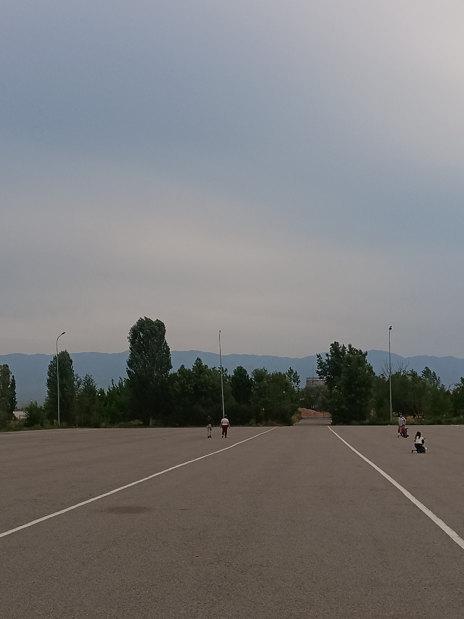 An asphalt parking lot is depicted with two white lines. The lot stretches towards a line of trees and distant blue mountains under a hazy, pale sky. Two individuals are walking on the far right side of the lot. In the mid-ground, two children are riding what appear to be scooters or similar wheeled devices. One child is wearing a blue shirt and dark shorts, and the other is wearing a white shirt and dark pants. A third child, in the middle distance, is also on a wheeled device, wearing a light-colored shirt and dark shorts. The environment suggests an outdoor recreational area or an open space near a town or village.