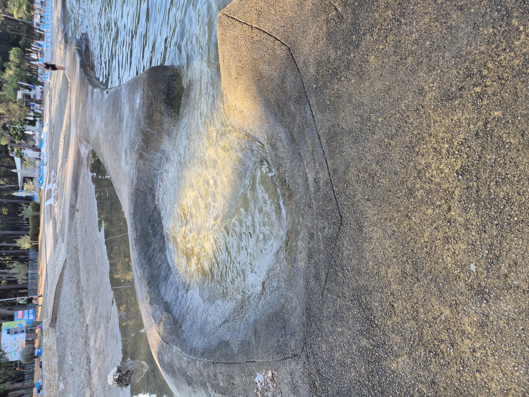 A concrete structure, resembling a pier or walkway, extends into a body of water. A circular depression in the concrete structure is filled with water, creating a rippling effect due to the movement of the waves. The water is clear and reflects the sunlight, casting a warm glow on the scene. In the background, a sandy beach is visible, lined with trees and umbrellas. A few lounge chairs are scattered on the sand, and some people are seen walking and relaxing. The overall scene depicts a tranquil beach setting on a sunny day, likely in the city of Monterol, Croatia.