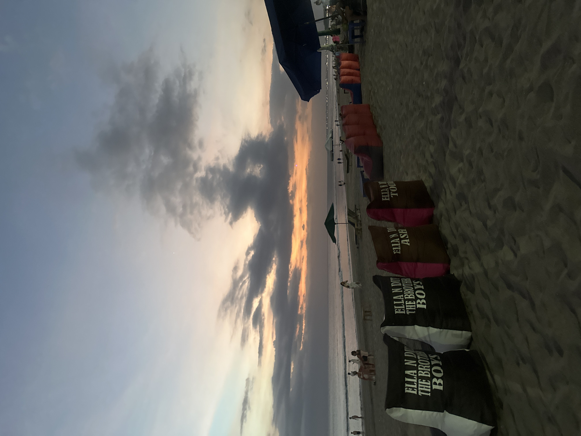 The image shows a beach scene, likely in Kuta, Indonesia, given the "ELLA N DU" branded cushions. The scene features rows of beach loungers and umbrellas stretching along the sand. There are beachgoers visible near the water's edge. The sky is filled with dramatic, dark clouds tinged with orange from the sunset. In the foreground are several cushions with various names printed on them. The sand appears to be dark.