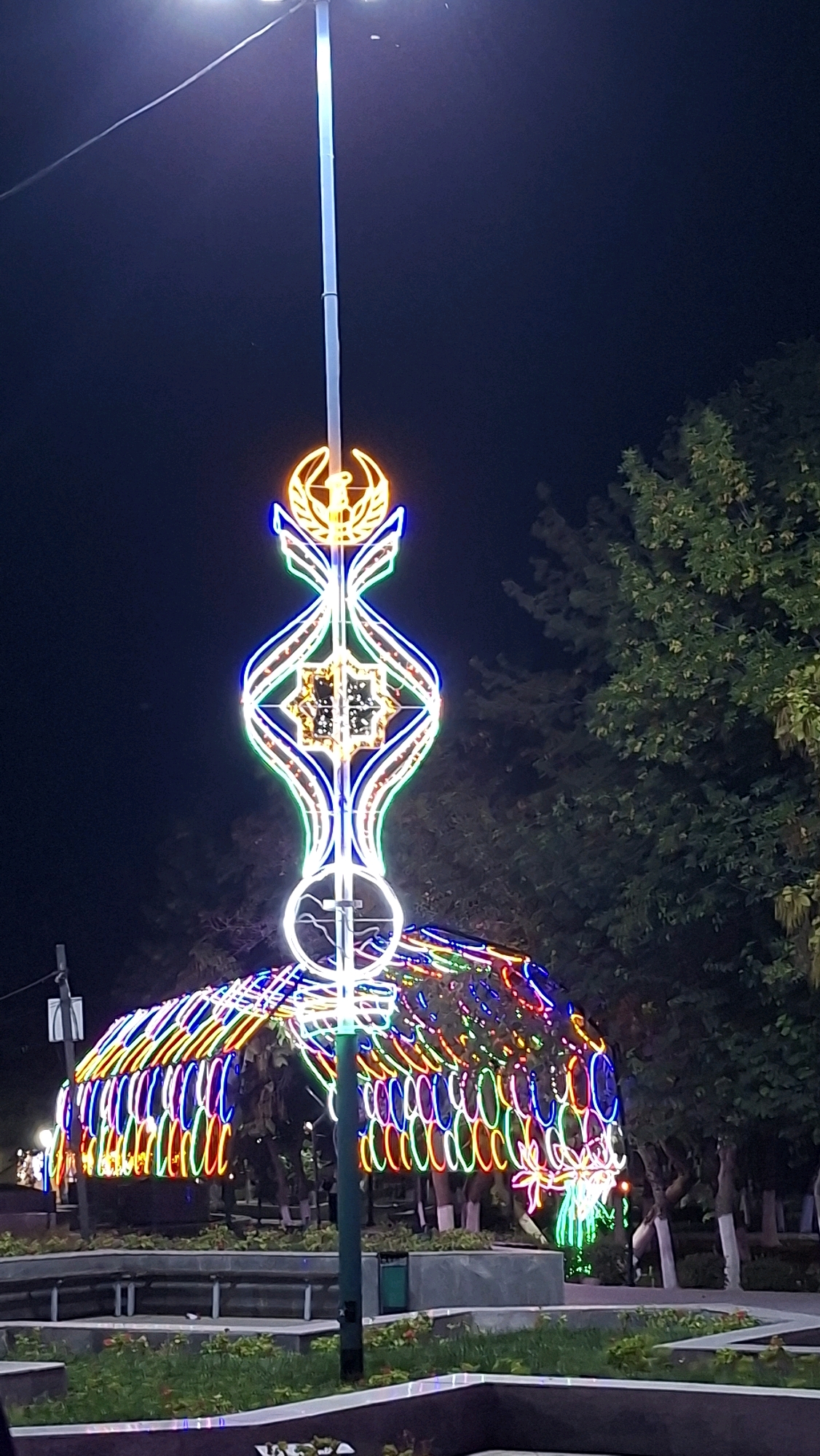 A vertical, full-frame, outdoor, night-time photograph captures a decorative light installation in Navoiy City, Uzbekistan. The installation is centered around a lamppost, which has a large, illuminated neon-like structure extending upwards and outwards from its middle section. This structure is symmetrical, featuring intricate, geometric patterns and a crest-like emblem at its apex, all outlined in various colors of light, including yellow, blue, green, and white. Below this, a larger, arched structure is also covered in vibrant, multicolored lights, forming a tunnel-like effect. The background is dark, indicating nighttime, with the silhouettes of trees visible to the right. In the foreground, there are low-lying planters with some greenery and benches. The scene appears to be a public space, possibly a park or square, adorned for an event or display. There are no people visible in the frame.