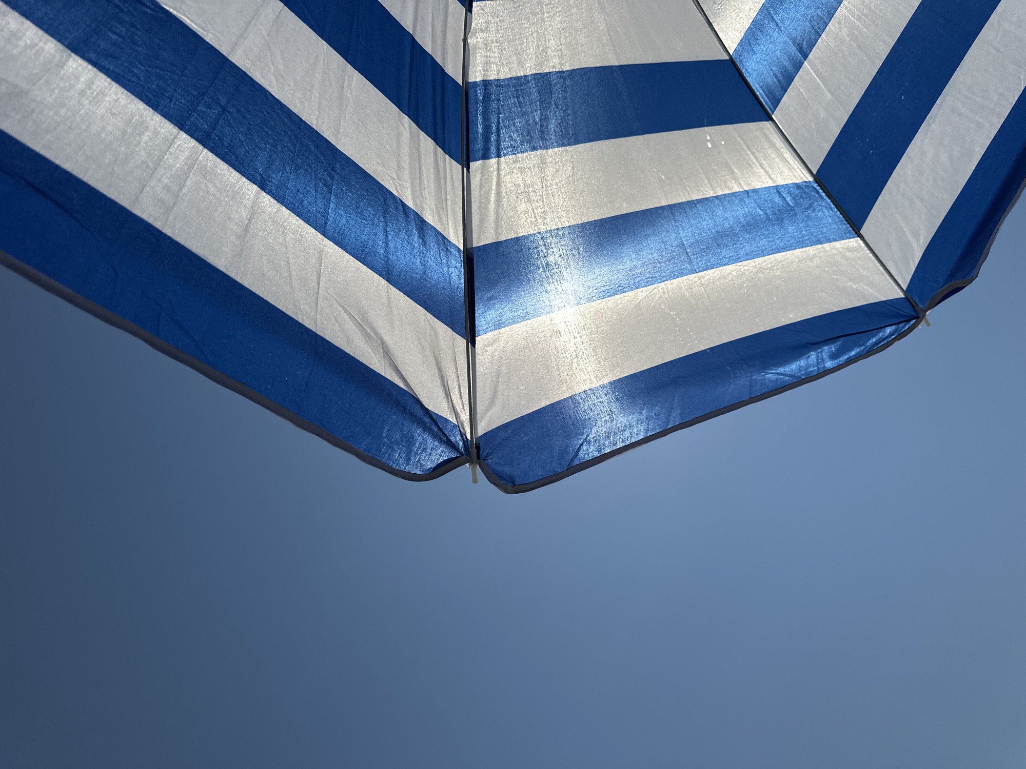 An upward-facing shot captures a blue and white striped beach umbrella against a clear blue sky. The umbrella occupies the right and bottom portions of the frame, its striped fabric angled diagonally. Sunlight filters through the fabric, creating bright highlights on the white stripes. The left side of the image is dominated by the expanse of the sky, devoid of any other objects or features. The setting appears to be a beach in Konyaaltı, Turkey. There are no people visible in the image.