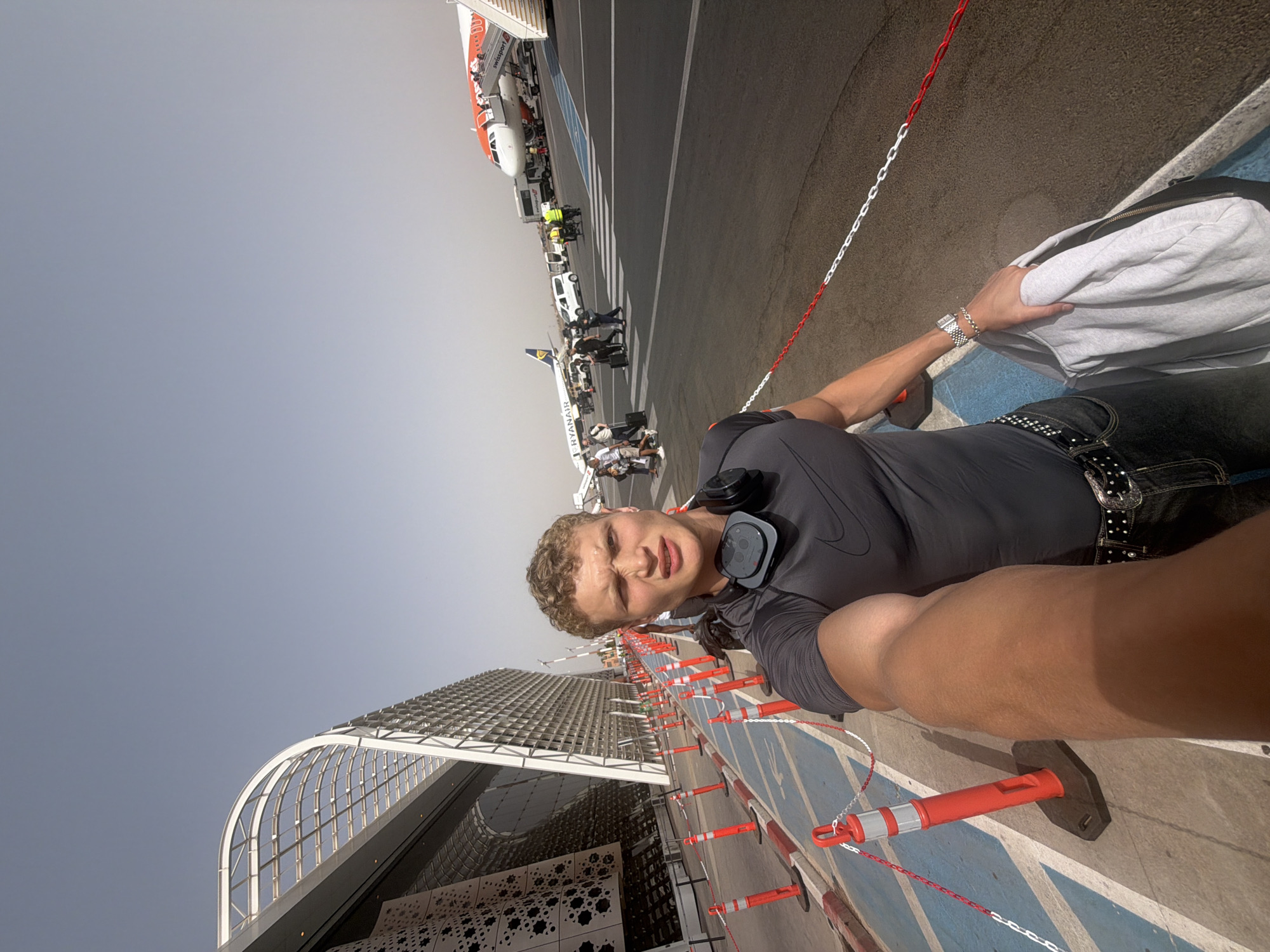 The image shows a young man with blonde, curly hair and headphones around his neck, taking a selfie in an airport. He is wearing a dark grey t-shirt with a Nike logo and dark jeans. He is holding a light grey jacket over his arm. The background shows an airport tarmac with several airplanes, including a Ryanair plane and an easyJet plane, as well as a modern airport building with a curved glass roof. People are seen walking on the tarmac, and there are several orange safety cones and barriers lining a pathway. The sky is clear and blue, indicating a sunny day. The architecture of the airport building suggests it is Marrakech Menara Airport, known for its modern design.