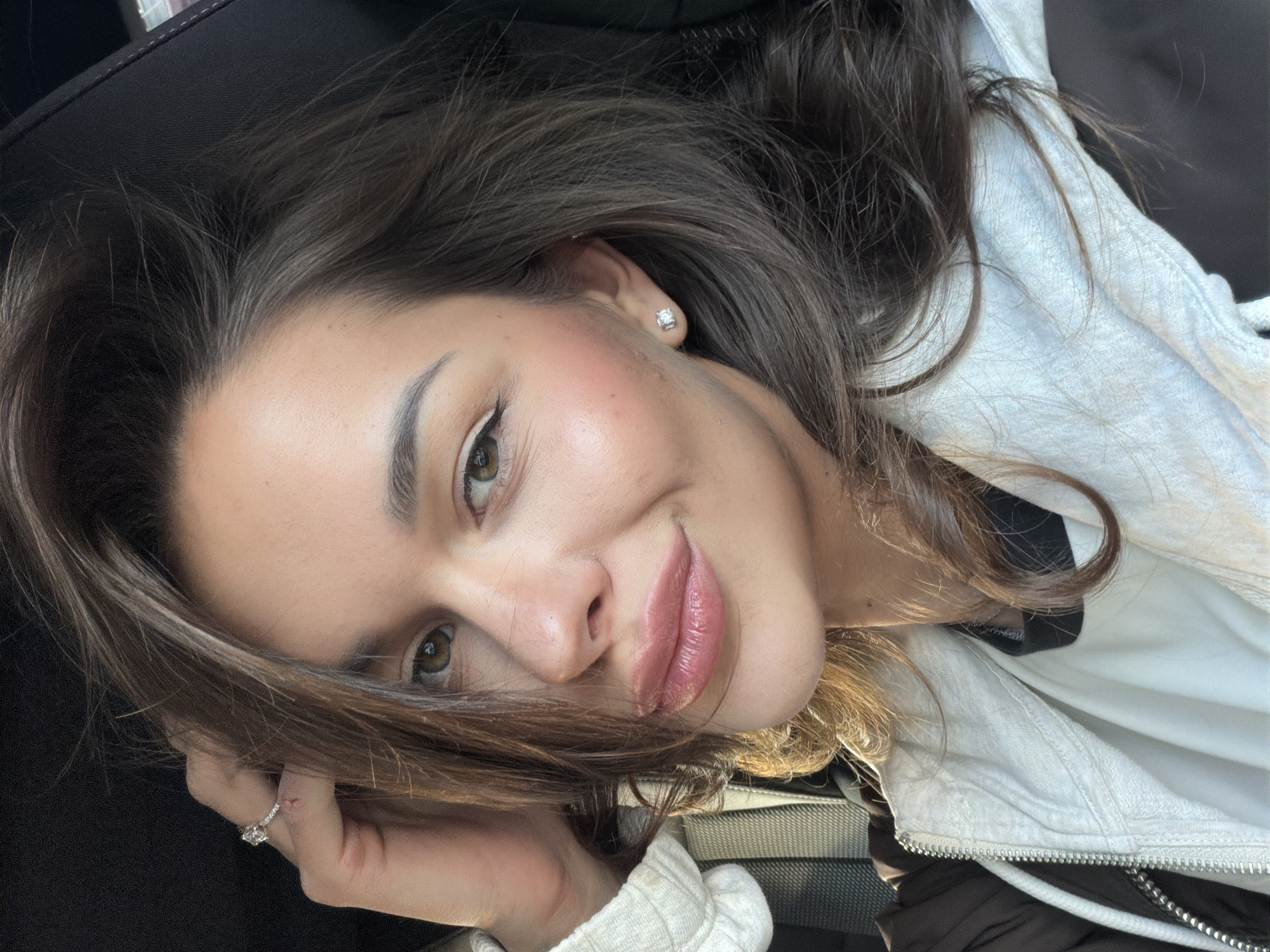 A headshot-style portrait captures a woman with long, dark brown hair, tilted to the right, suggesting she is lying down or has taken a selfie from a low angle. She looks directly at the camera with a gentle, soft smile. Her light brown or hazel eyes are accented with subtle eyeliner and mascara, complementing her well-defined dark eyebrows. Her full lips are adorned with a glossy pink or nude lipstick, and a hint of blush is visible on her cheeks.
She wears a small, square diamond stud earring in her right ear. On her left ring finger, a prominent diamond ring with a central stone and smaller diamonds on the band is visible, held close to her face with her fingers partially obscured by her hair. She is dressed in a light-colored, possibly white or off-white, hooded jacket or sweatshirt, worn over a darker garment, elements of which are seen around her neck and shoulder. The background appears to be the dark, upholstered interior of a vehicle, likely a car seat, providing a simple, unfocused setting. The lighting is soft, indicative of daytime. There is no visible text in the image. The provided context indicates the location is Krasnoyarsk, Russia.