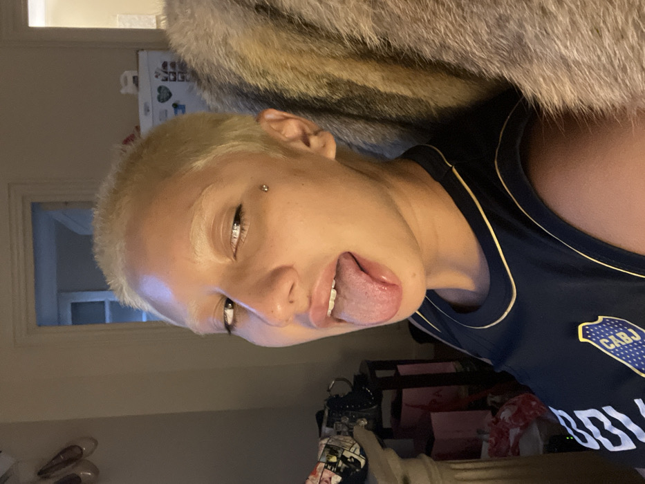 An individual with bleached blonde short hair and eyebrows, featuring a dermal piercing on the left temple, is captured in a close-up indoor portrait. Their mouth is open, with the tongue extended. They are wearing a dark blue sleeveless jersey with yellow piping and the official shield logo of Club Atlético Boca Juniors, including the "CABJ" acronym. The background elements include a light-colored wall with two hanging light-toned ballet flats and a dark bag on a surface to the left. A doorway is visible centrally. To the right, a white refrigerator with a heart-shaped magnet and a large, textured light-brown/grey fur-like object are present. The environment appears to be a residential interior under artificial illumination. The Boca Juniors jersey identifies the location as culturally situated within Buenos Aires, Argentina.