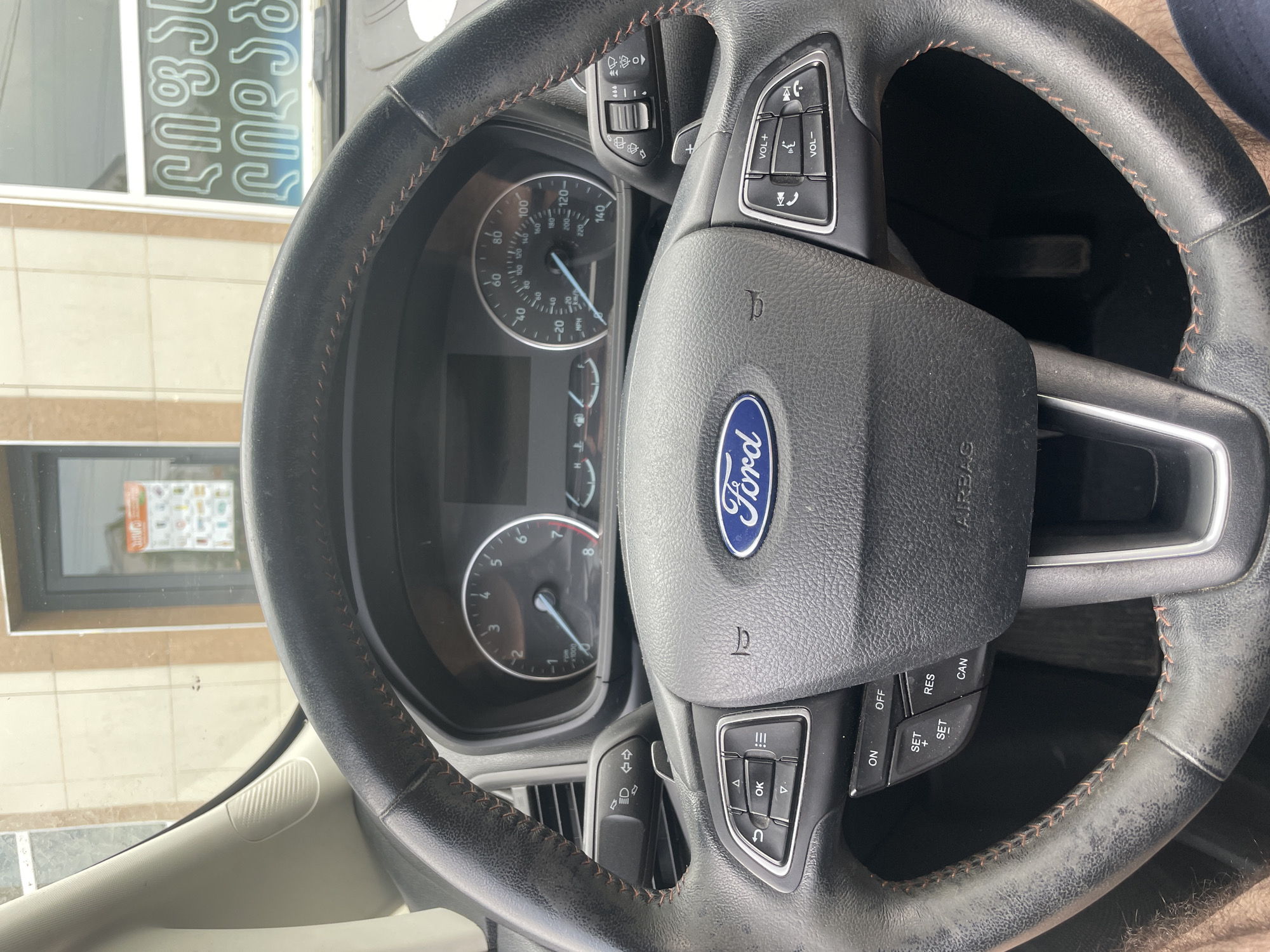The image shows the interior of a Ford vehicle, focusing on the steering wheel and dashboard. The steering wheel is black with orange stitching and has controls for volume, calls, and media on the right side. On the left side, there are controls for navigation, settings, and trip information. The dashboard features a speedometer, tachometer, and fuel gauge. The Ford logo is prominently displayed in the center of the steering wheel. Outside the vehicle, visible through the windshield, is a building with windows and signage in a foreign script, suggesting the car is parked in a city. The overall lighting indicates it is daytime.