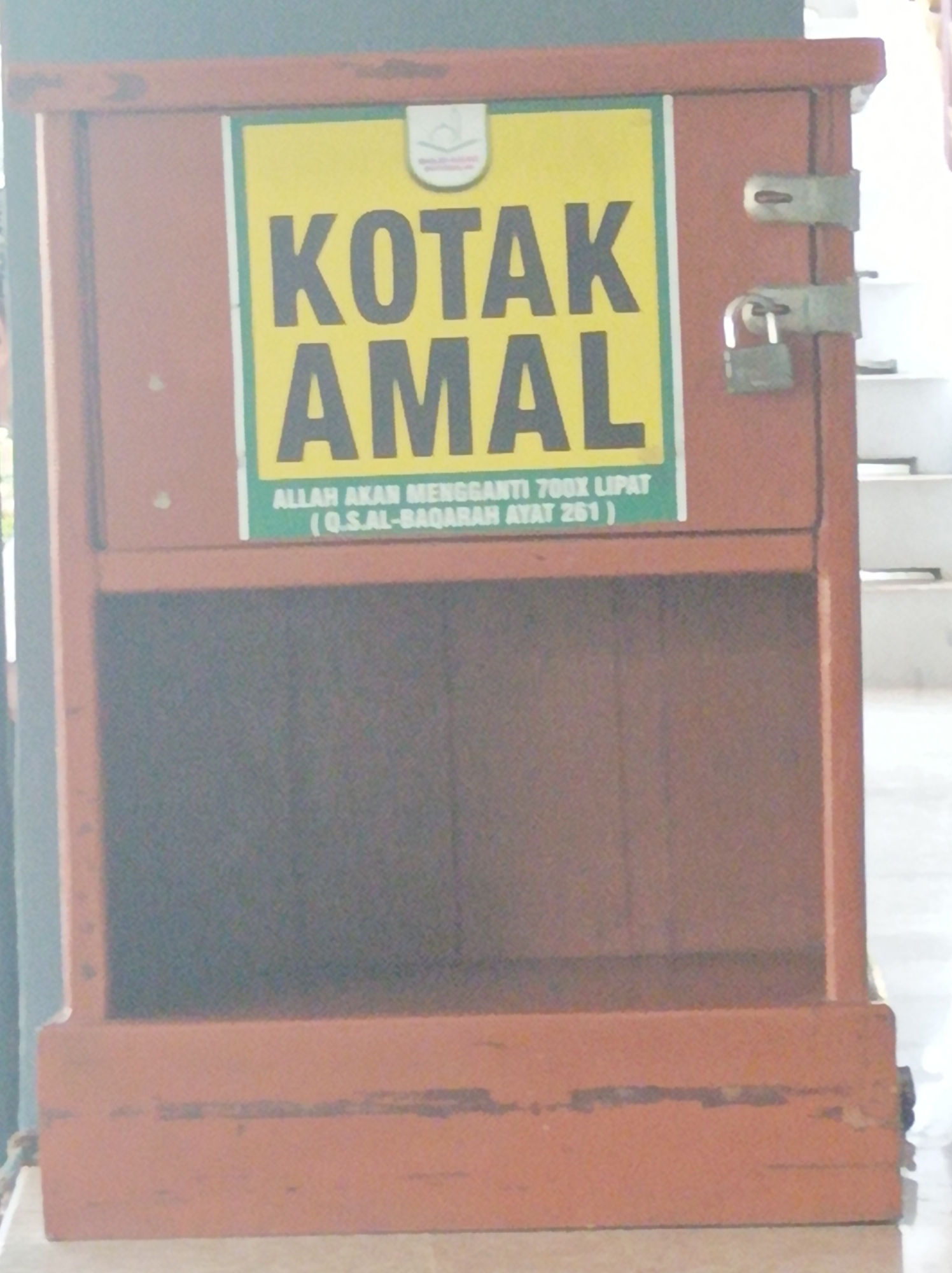 The image shows a wooden donation box, labeled "KOTAK AMAL" in large black letters on a yellow background. Below this, smaller text reads "ALLAH AKAN MENGGANTI 700X LIPAT (Q.S.AL-BAQARAH AYAT 261)". The box is orange-brown and has two metal latches and one padlock securing its opening. The background suggests an indoor environment with some shelves visible. The image is a medium shot, focusing on the donation box. Based on the signage and the context of donation boxes often found in public places in Indonesia, this donation box is likely located in the city of Nganjuk, Indonesia, though its specific location within the city is not discernible from the image.