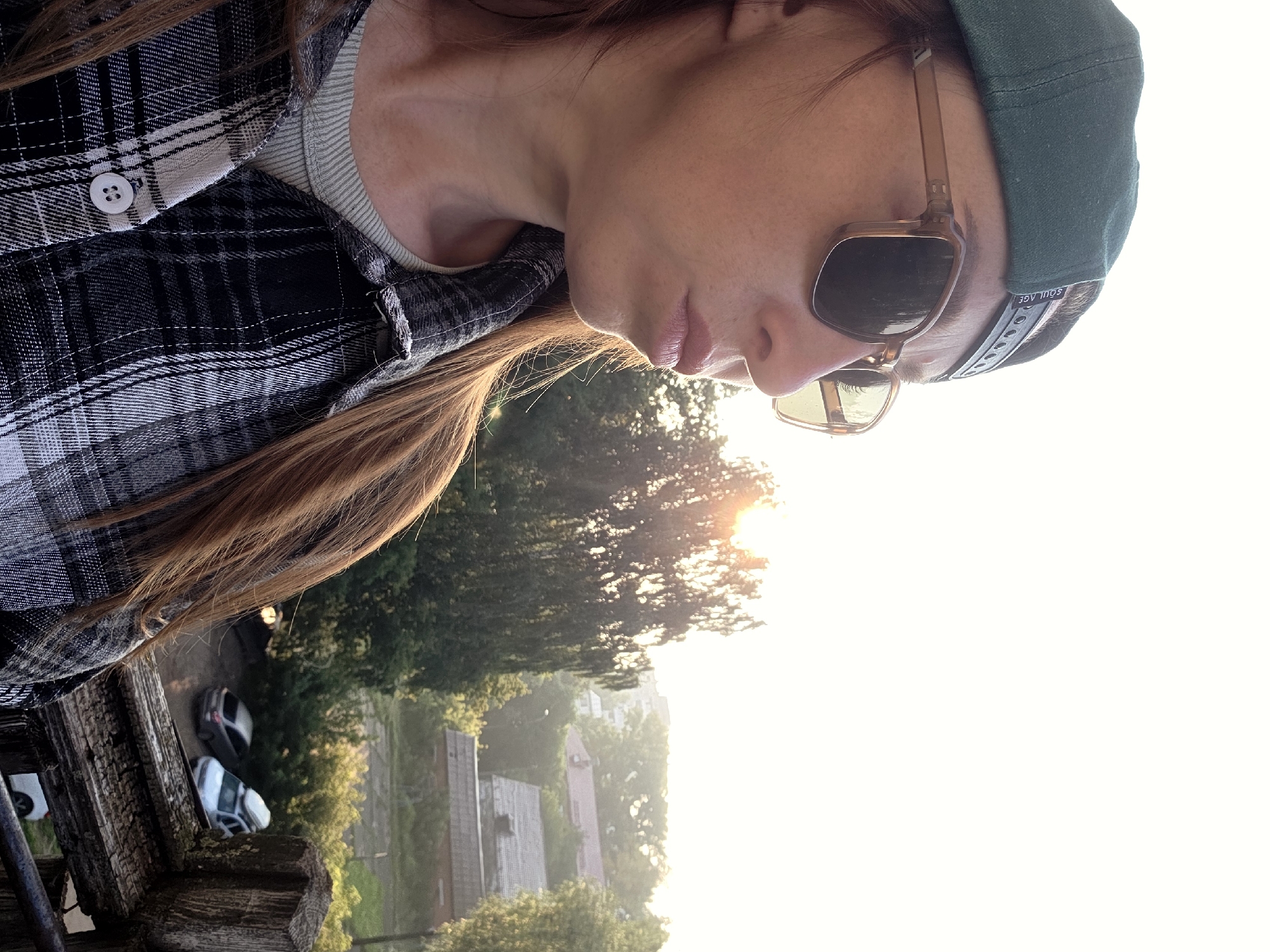 A young woman with long, auburn hair is captured in a close-up, profile shot, her face turned downwards and away from the camera. She is wearing a black and white plaid shirt over a light-colored t-shirt, and a dark green baseball cap. Her face is partially obscured by a pair of square-framed sunglasses with amber lenses. The lighting suggests it is late afternoon or early evening, with the sun low in the sky, casting a warm glow and creating a lens flare effect that illuminates the background.
The background reveals a hazy, semi-rural scene. Trees with lush green foliage are visible, interspersed with rooftops of houses, suggesting a residential area. In the foreground, a weathered wooden railing or fence runs along the bottom left of the frame. Two cars are parked on a dirt or gravel surface behind the railing. The overall impression is one of a casual, outdoor moment, possibly taken on a balcony or porch overlooking a neighborhood. The woman's expression is unreadable due to her lowered gaze and sunglasses, but the overall mood is serene and relaxed. The visible text on the back of the baseball cap appears to be "SOUL AGE". The image has been rotated, with the sky appearing at the right of the frame.