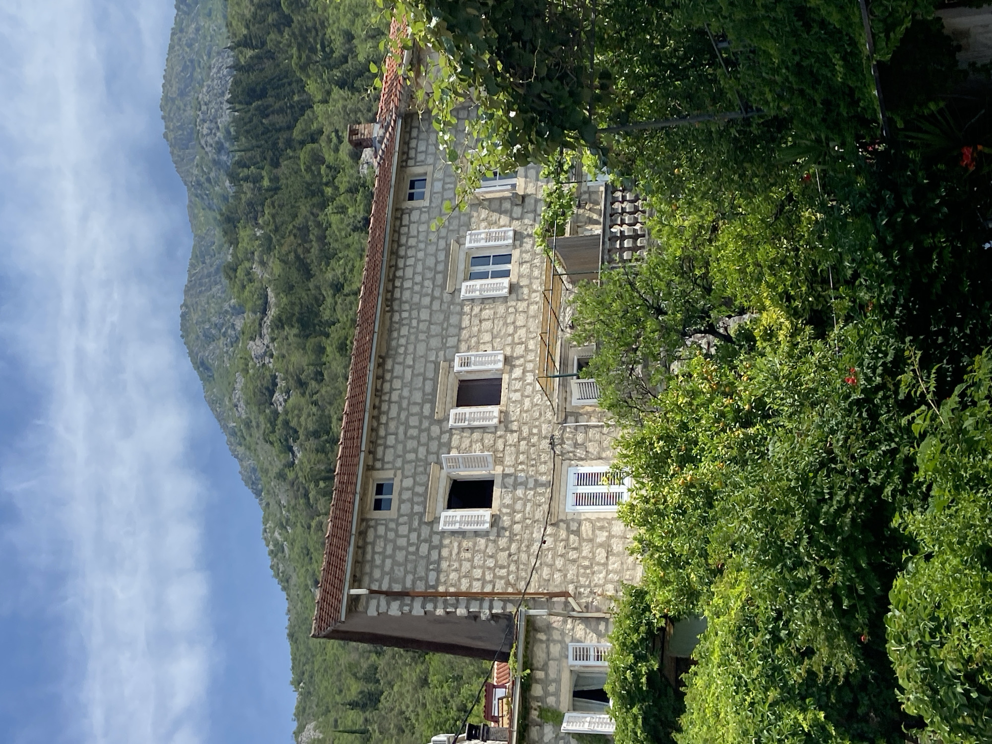 The image shows a stone building with a red tile roof, nestled against a lush green mountain backdrop in Perast, Montenegro. The building has multiple windows with white shutters, some of which are open, revealing dark interiors. A balcony with an ornate railing is visible on the right side of the building. Abundant green foliage, including trees and vines, surrounds the lower portion of the building, partially obscuring it from view. Some red flowers are also visible amongst the greenery. The sky above is blue with scattered white clouds. The scene depicts a residential building in a mountainous coastal town.