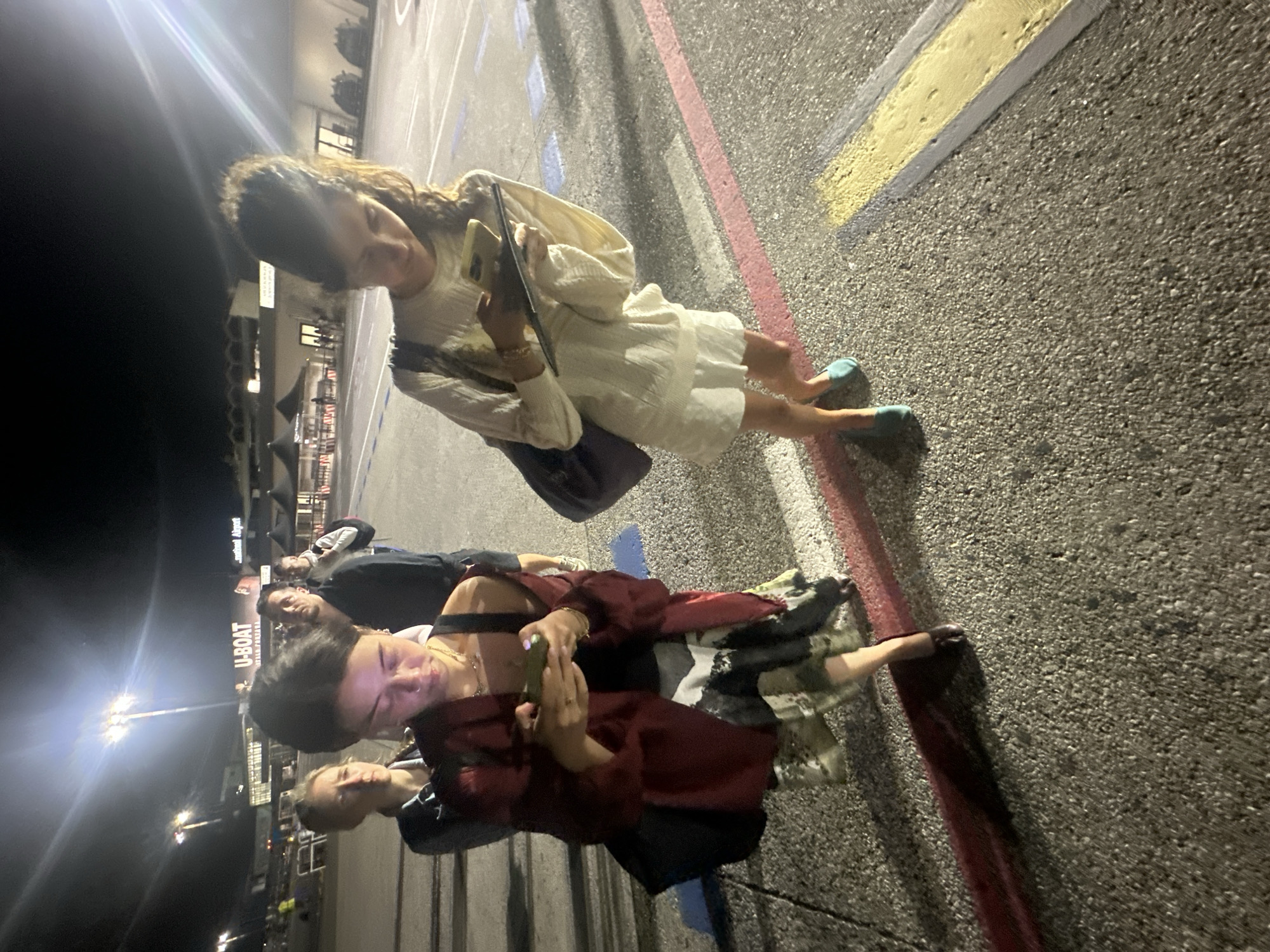 An outdoor night scene captures a group of young individuals gathered on a paved surface, likely at an airport or a large public area in Pisa, Italy. The image is tilted, giving a dynamic perspective.
In the foreground, a woman with long, dark, wavy hair wears a light-colored, possibly white or cream, dress and light blue flat shoes. She holds a smartphone in one hand and what appears to be a tablet or book in the other, looking down at her devices. A dark shoulder bag is slung across her body.
Behind her and to the left, several other people are visible. One person in a dark red jacket and a patterned skirt or dress is also engaged with a smartphone. Another individual with lighter hair stands nearby. Further back, two more people in dark clothing are present, with one looking towards the camera.
The ground is asphalt, marked with painted lines in red, yellow, blue, and white, resembling parking lot or pathway markings. Bright artificial lights from street lamps illuminate the scene, creating significant lens flares and casting shadows, indicating it is nighttime.
In the background, buildings are visible. A prominent sign clearly displays "U-BOAT" in large letters, with "ITAN.PORADA" in smaller text beneath it. Another partially visible sign further back reads "tinental Airport," strongly suggesting the location is Pisa International Airport.
The individuals appear to be casually waiting or gathered, with several occupied by their mobile devices. The overall mood is relaxed and casual.