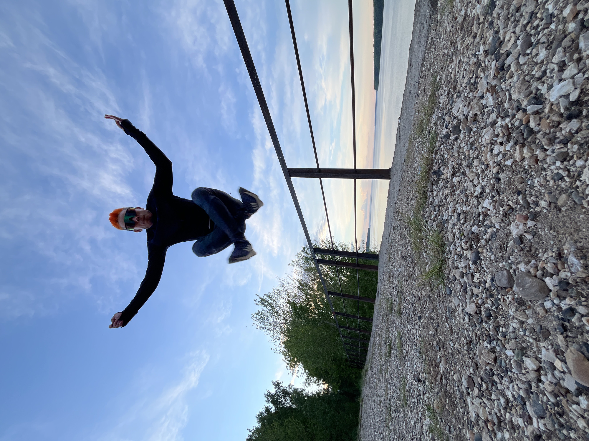 A man jumps in the air near a metal railing. He has red hair, wears dark clothes, and orange-tinted sunglasses. The setting appears to be outdoors, next to a body of water with a cloudy sky. Vegetation is visible in the background. The ground is covered in pebbles. The image is from the city of Chaikovsky, Russia.