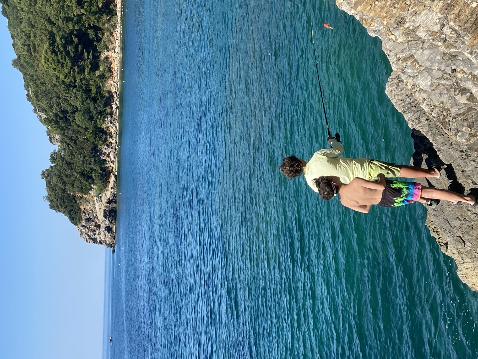 Two boys are fishing on a rocky coastline overlooking a clear turquoise sea. The boy in the foreground is wearing colorful swim trunks and has his back to the camera. The boy behind him is wearing a yellow long-sleeved shirt and shorts. They are both holding fishing rods, with one rod in the water. In the background, a lush green cliff juts out into the sea, with a small boat visible in the distance. The sky is a clear blue. This scene is characteristic of the coastline near Budva, Montenegro, known for its beautiful beaches and rocky coves.