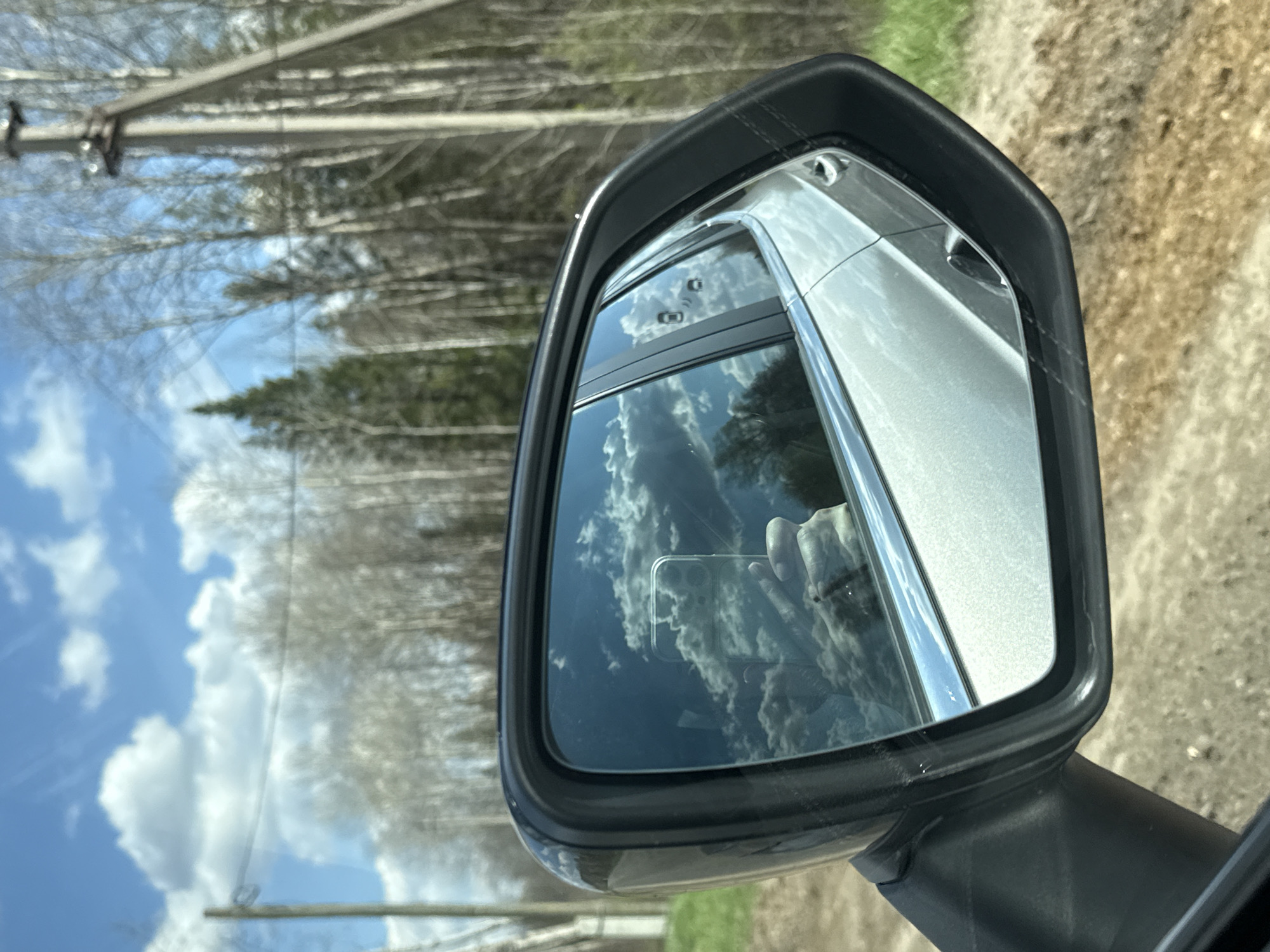 The image shows a car's side mirror reflecting a view of the sky with scattered clouds and trees. The reflection also captures the photographer's hand holding a smartphone. Outside of the mirror, a dirt road and power lines are visible. The location cannot be confirmed as Бикбарда, Russia based on the provided image.