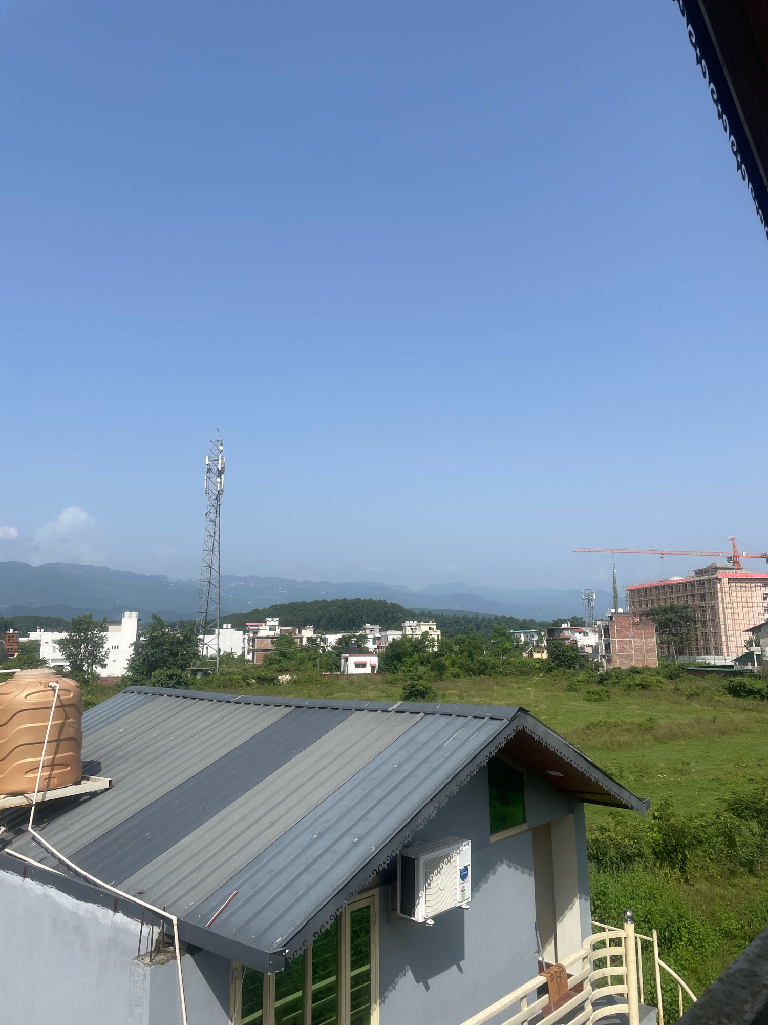 The image shows a view of Kolhupani, India, on a clear day. In the foreground, there is a gray building with a metal roof that has alternating dark gray and lighter gray stripes. A large, tan water tank sits on the roof of the building on the left. An air conditioning unit is mounted on the side of the building on the right.
In the mid-ground, there is a collection of buildings, including a tall cellular tower. Further in the distance, there are more residential buildings and some green trees. On the far right of the mid-ground, a multi-story building under construction is visible, along with a red construction crane.
The background consists of rolling, green hills and mountains under a bright blue sky. The overall scene depicts a semi-urban or suburban environment with evidence of ongoing development.