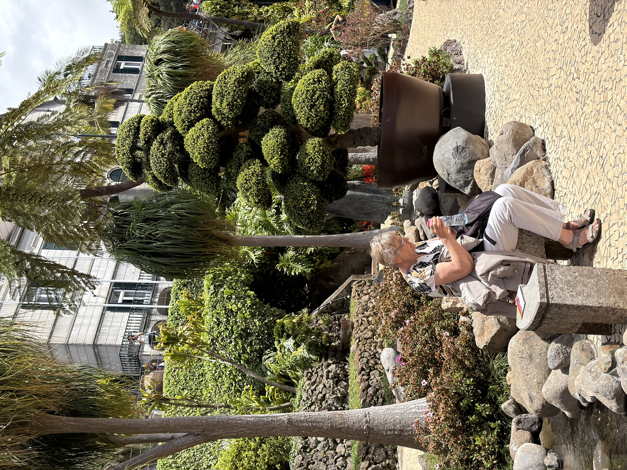 A woman sits on a stone bench in a verdant garden setting, holding a water bottle. The scene is likely in the city of Funchal, Portugal, as indicated by the lush vegetation and architectural style. The garden features a variety of plants, including meticulously shaped trees, palm trees, and flowering shrubs. The woman wears light-colored pants and a patterned shirt. A stone pathway runs behind her. A grand building is visible in the background, partly obscured by the foliage.