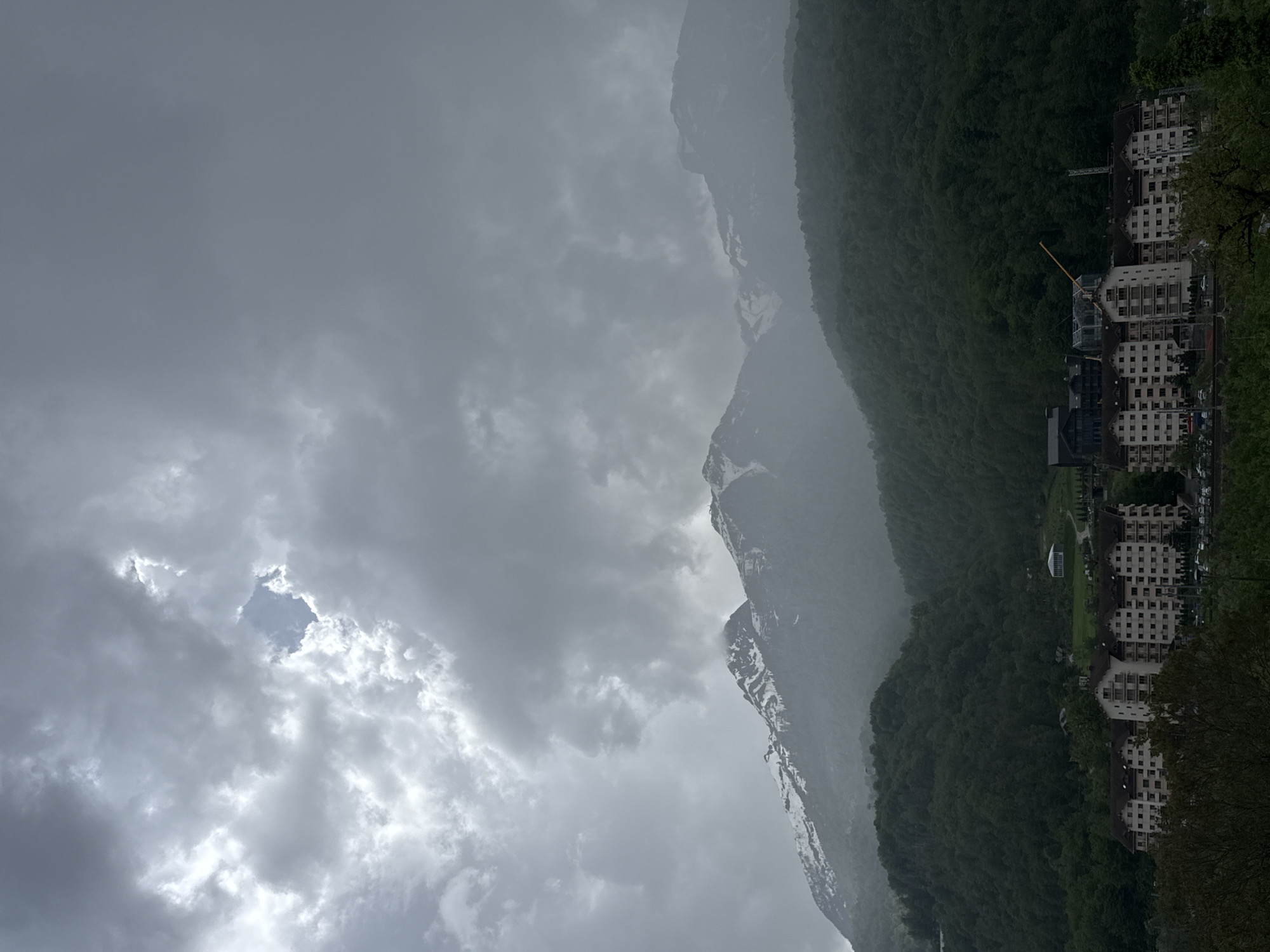 The image depicts a landscape scene in Krasnaya Polyana, Russia, on a cloudy day. In the foreground, a range of mountains, some with snow-capped peaks, is visible. Dense green forest extends along the base of the mountains, adjacent to buildings resembling hotels or apartments. The sky is overcast with thick, layered clouds, revealing a small patch of blue sky. A construction crane is visible near the buildings. The overall ambiance is gray and somewhat somber.