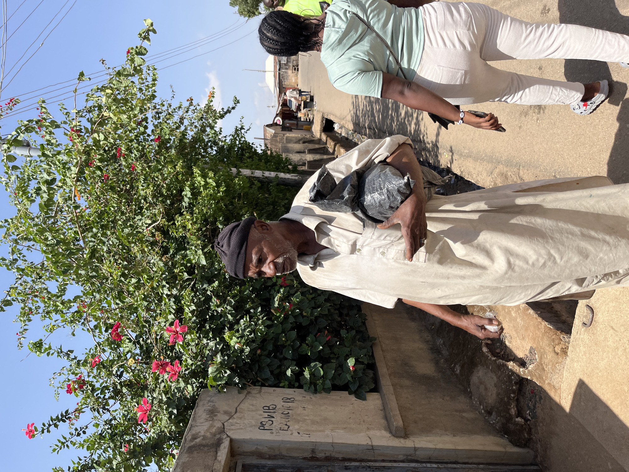 An elderly man with a black knit cap is standing on a street in Jos, Nigeria. He is carrying a black plastic bag in his left arm. He wears a light tan, long-sleeved shirt. Beside him is a concrete structure with painted graffiti and a flowering bush with red flowers. The ground has an uncovered drainage ditch. Further away, a woman in a green shirt and white pants walks along the street. In the background are other people and buildings.