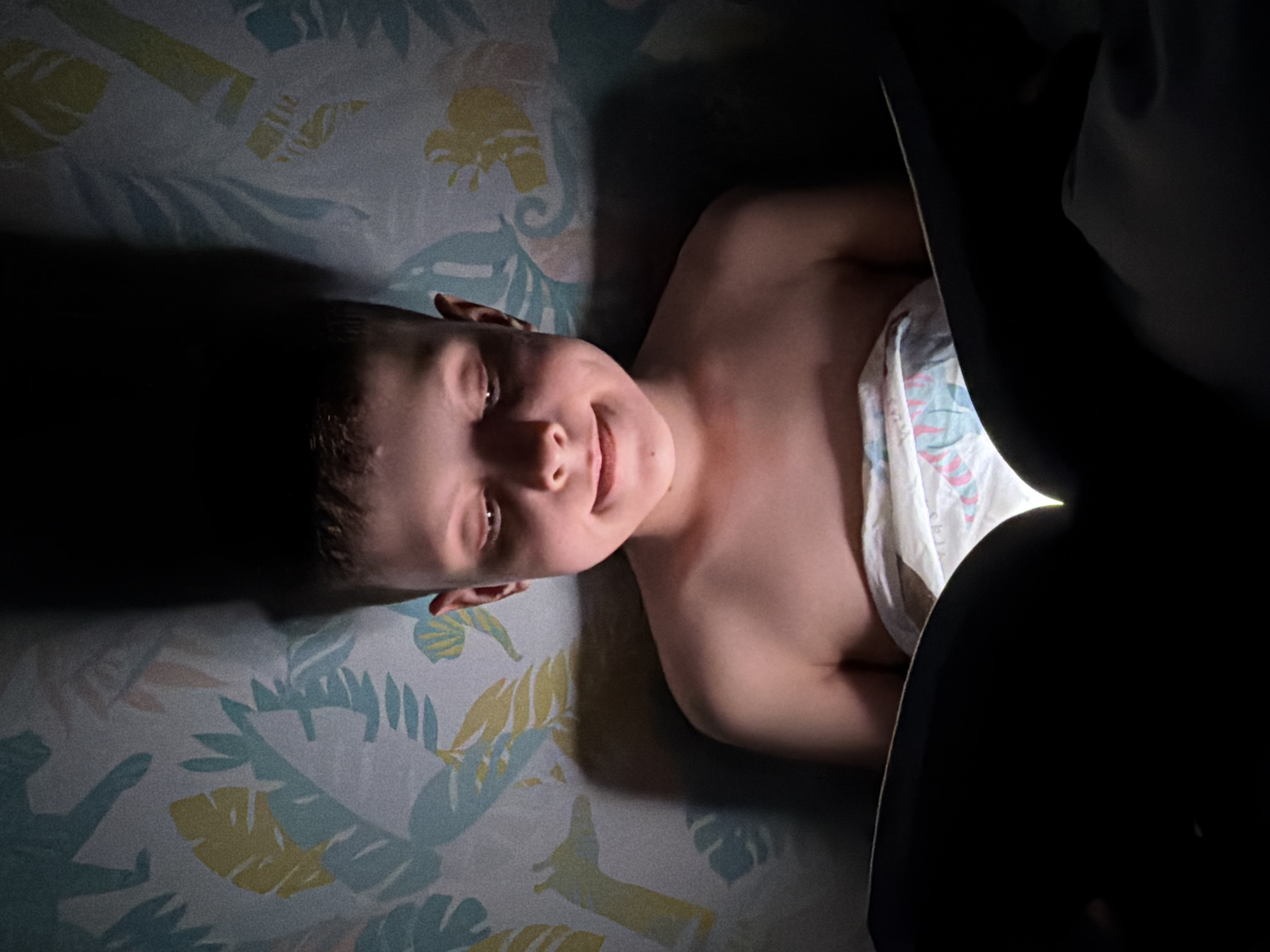 A young, bare-chested child lies on a bed with a patterned sheet, partially obscured by a dark object in the foreground. The child is smiling and looking toward the camera. No information is available about the location of this scene in Votkinsk, Russia.