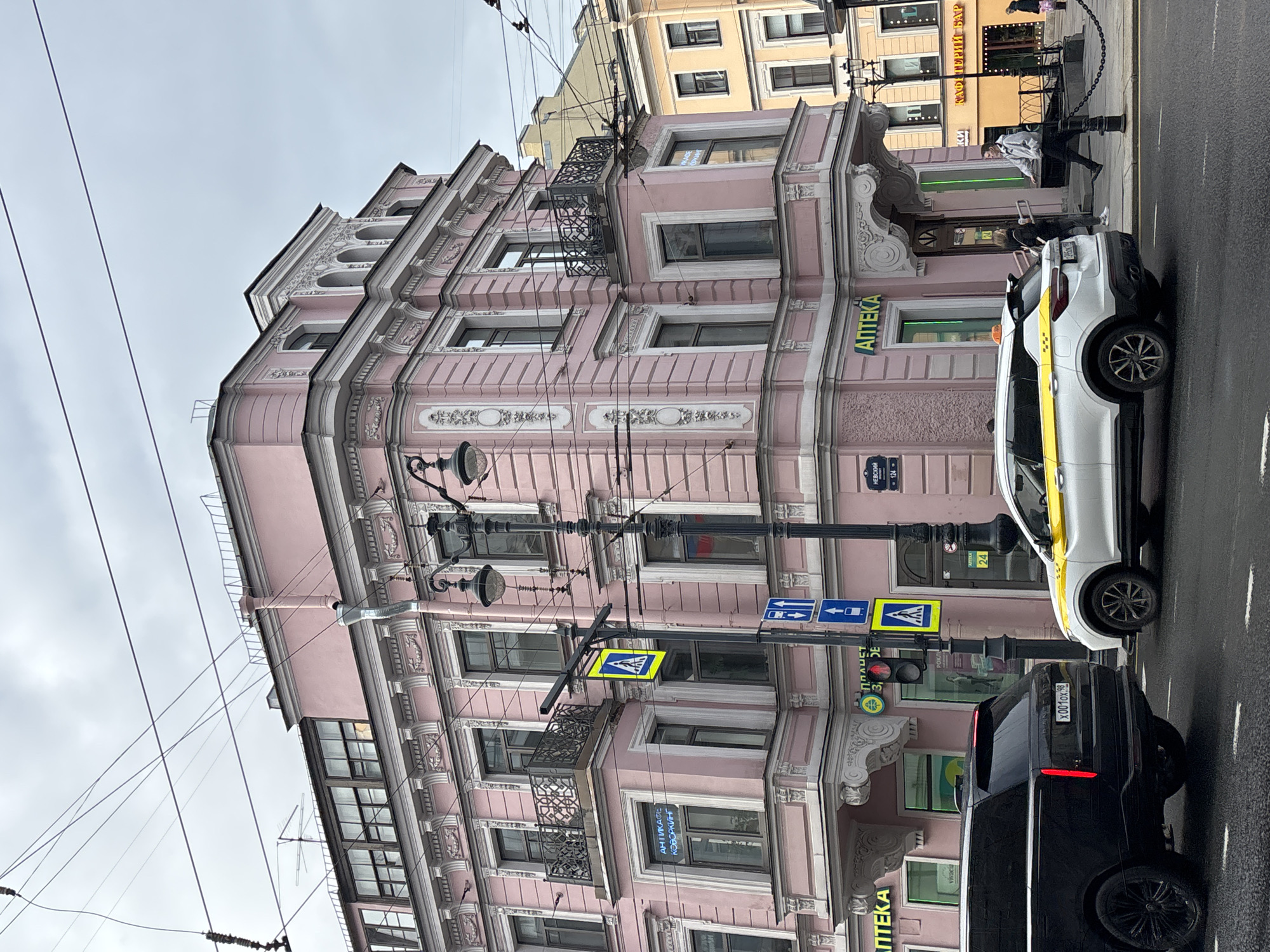 The image depicts a pink, multi-story building in St. Petersburg, Russia, identifiable by the Russian signage and architectural style. Street signs in Russian and crosswalk signals indicate a pedestrian crossing. A white taxi with a yellow stripe and a black SUV are parked on the street in front of the building. There are pedestrians on the sidewalk. "Aптека" (Pharmacy in Russian) signage is visible on the building's facade. Overhead are power lines and light poles. A street sign on the corner says "Невский" (Nevsky), which means Nevsky prospect.