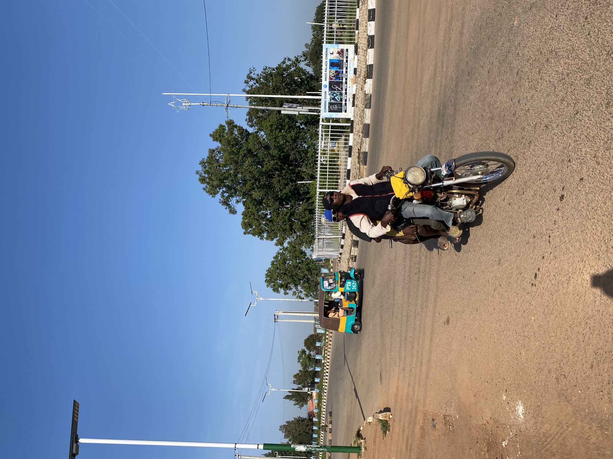 In Bar Jirgi Summa, Nigeria, a sunny day unfolds on a paved road, the sky a clear, vibrant blue. A man, wearing a black vest over a white shirt and blue jeans, rides a red motorcycle, a yellow bag secured to the handlebars. He is accompanied by another person seated behind him. To their left, a teal and yellow tuk-tuk, with the license plate PTA-3634, transports three passengers.
The road is bordered by a white metal fence, behind which trees and a large billboard advertising "MEES PALACE CINEMA" are visible. The billboard features images of movies and the text "COMING SOON." Streetlights with solar panels dot the landscape, and power lines stretch across the sky. The ground is a mix of paved road and dirt.