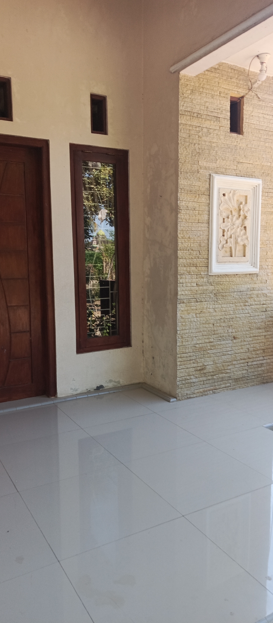 A building's exterior in Jatisari, Indonesia, features a beige wall with a wooden door on the left. Adjacent to the door is a narrow, vertical window with a dark wooden frame, reflecting trees and a dome structure outside. To the right, the wall transitions to a textured stone facade, adorned with a rectangular, white relief carving of flowers. Above this section, a small, square window is visible. The ground is covered in large, light-colored tiles, reflecting the surroundings. A white ceiling with a fluorescent light fixture and a fan is present at the top right.