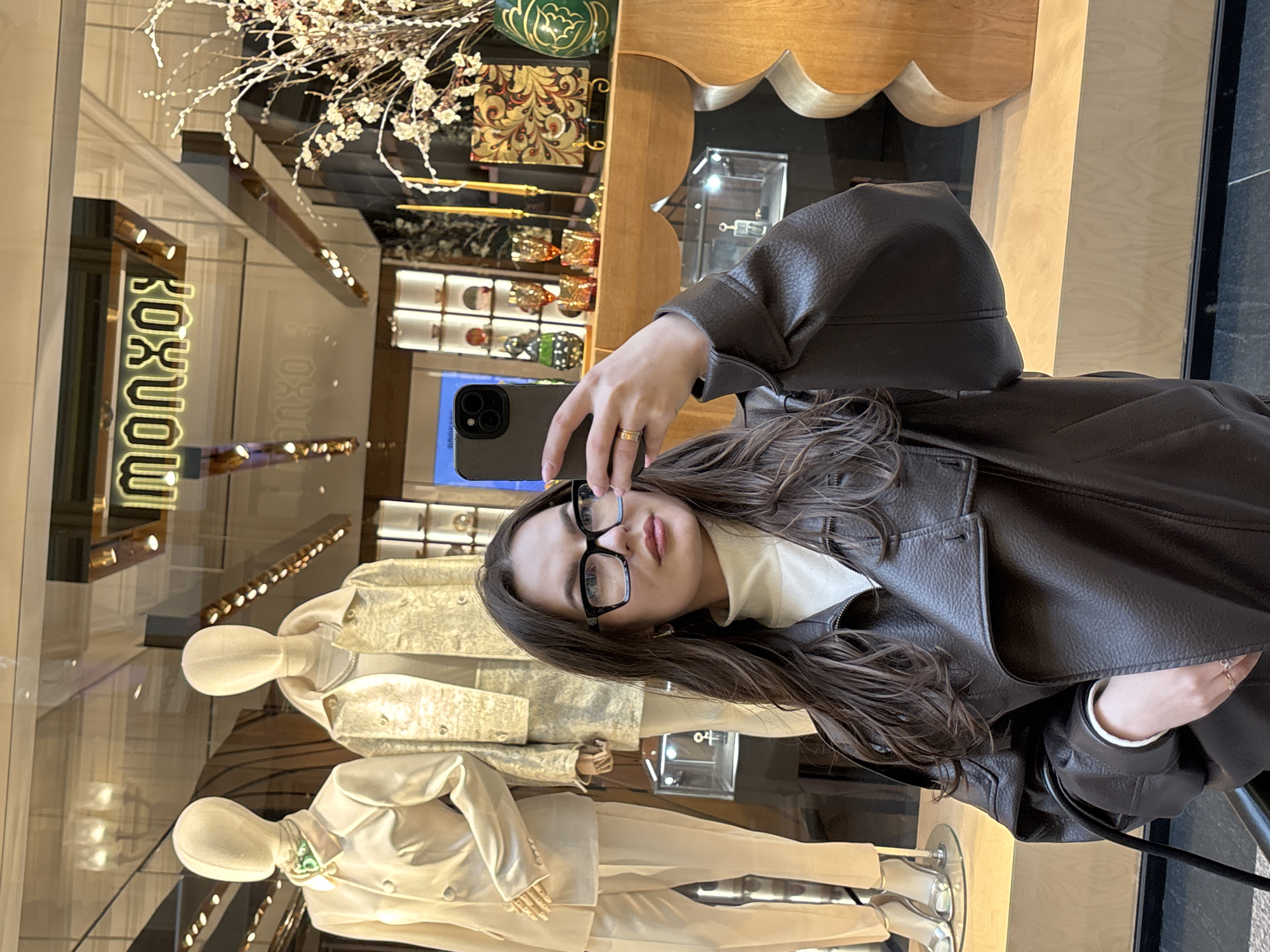 A woman with long brown hair and glasses is taking a selfie in front of a store window. The storefront has mannequins dressed in light-colored suits. Reflected in the glass is the store's signage, which reads "monxo." The store appears to be located in the city of Moscow, Russia, as identified by the signage and general visual style. The woman is wearing a brown leather jacket and holding her phone up to take the picture.