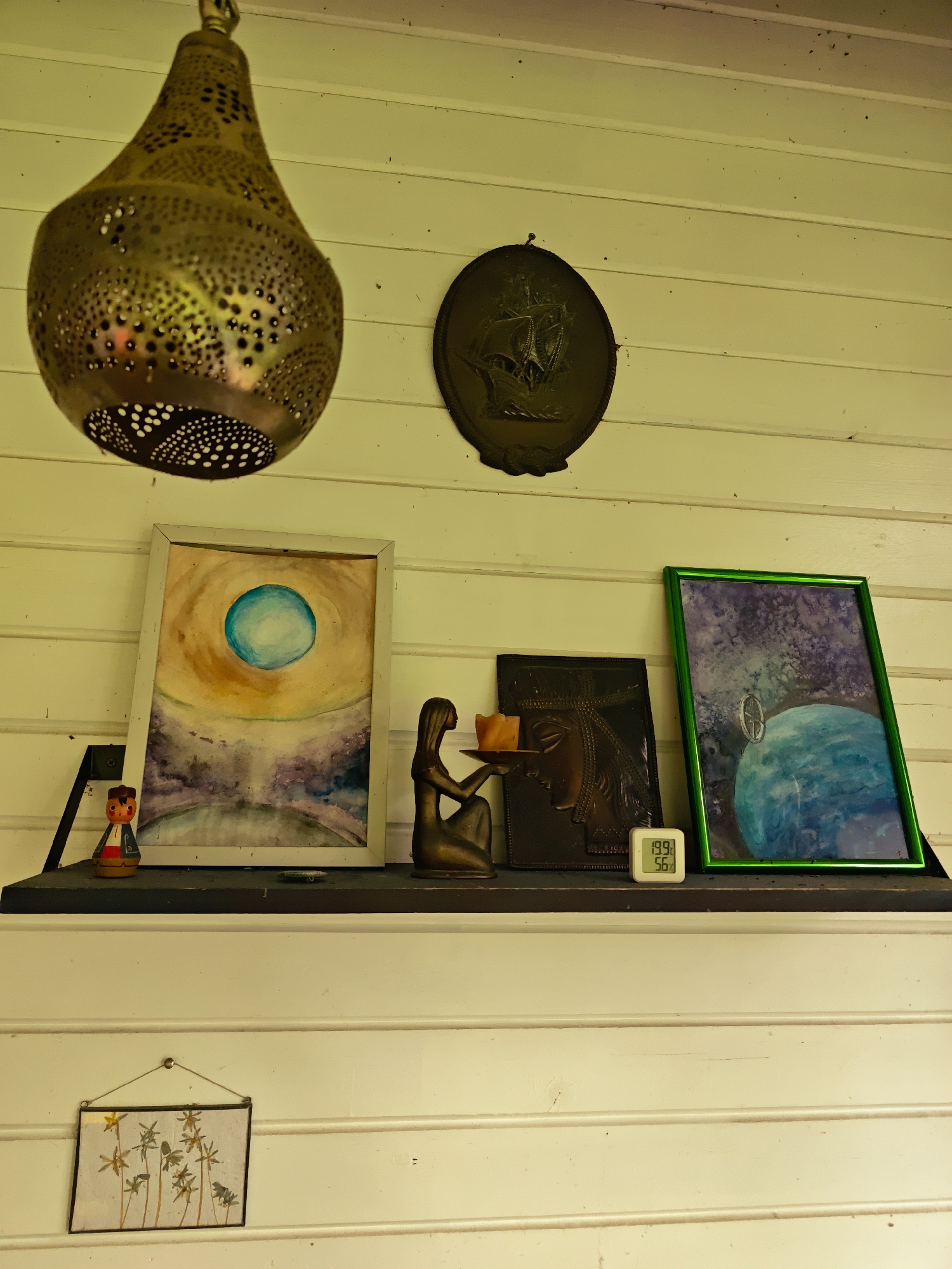 A still-life scene is captured against a white horizontal wooden panel wall. An ornate, gold-colored metal pendant lamp hangs from the ceiling, partially obscuring the upper-left corner. Adjacent to it on the wall is a circular, bronze-colored plaque depicting a sailing ship.
A black shelf is mounted against the wall, supporting an array of items. From left to right, there is a small, painted wooden doll figure; a framed watercolor painting with a white frame, depicting a teal-colored orb against a background of yellow and purple hues; a bronze-colored sculpture of a seated woman holding a dish aloft; a dark, rectangular piece, possibly a plaque or art panel depicting a stylized Egyptian face; and a small digital device displaying temperature and humidity readings, set next to a green-framed painting depicting an earth-like planet in a star-studded sky.
Hanging below the shelf, centered, is a small framed picture with a clear, transparent background, depicting stylized palm fronds. The overall lighting is soft, casting shadows and highlighting the textures of the objects and the wooden wall.