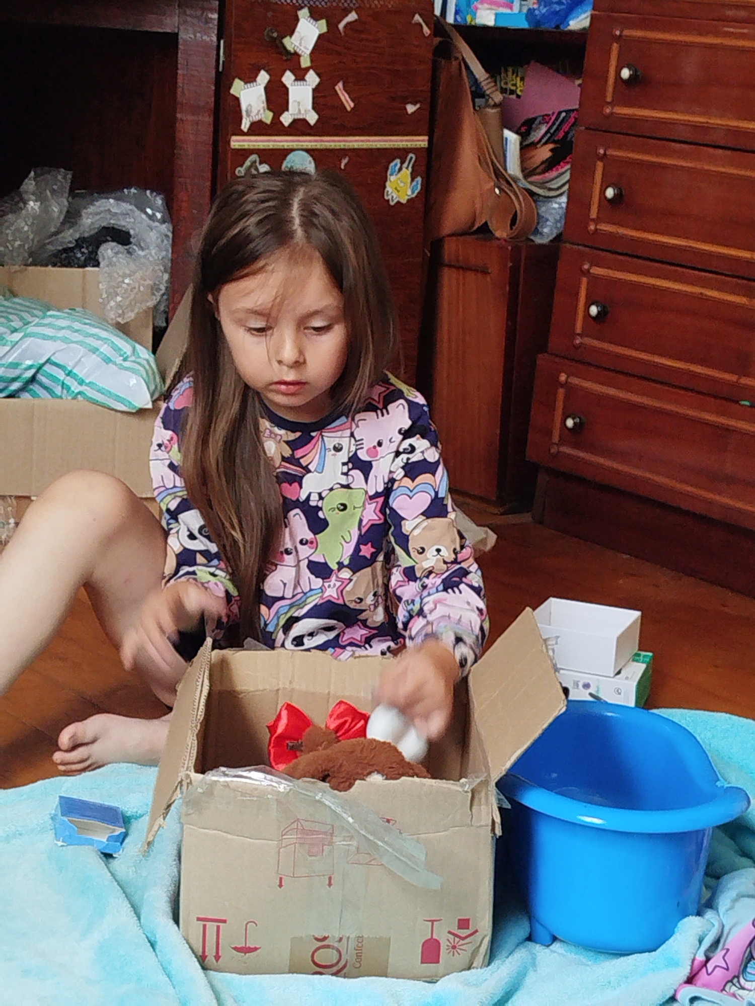 A young girl with long brown hair, wearing a long-sleeved shirt with cartoon animal prints, sits on a wooden floor. She is looking down at a cardboard box in front of her, which contains a brown stuffed animal with a red bow and some white objects. The girl's right hand is reaching into the box. To the right of the box is a bright blue plastic basin. The background consists of wooden furniture, including a dresser with drawers and shelves filled with items. The overall environment appears to be an indoor domestic setting, possibly a bedroom or living area. There are no specific visual cues to pinpoint the location to Vinnytsia, Ukraine.