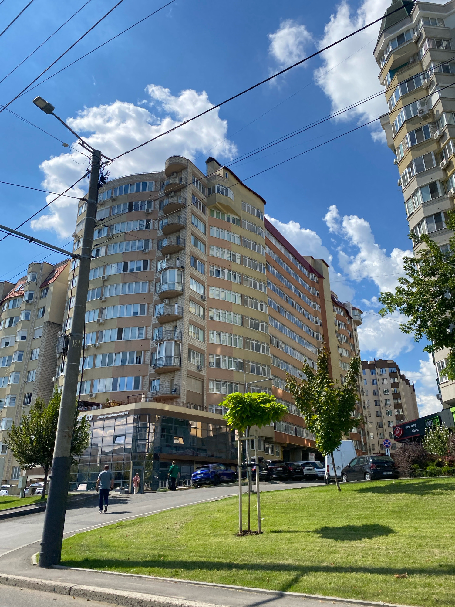 A low-angle outdoor shot captures a modern, multi-story building with a curved facade, featuring numerous windows and balconies, under a bright blue sky with scattered clouds. A lamppost stands in the foreground, to the left of the main building. Adjacent to it, a smaller glass-fronted building houses a sign that appears to say "co centre de réadaptation médical". Several cars are parked in front of the buildings, and two individuals are walking near the glass building. The scene is located in Chișinău, Moldova, as evidenced by the signage on the glass-fronted building which is a medical rehabilitation center. A green lawn with two young trees in the foreground leads up to the sidewalk and street.