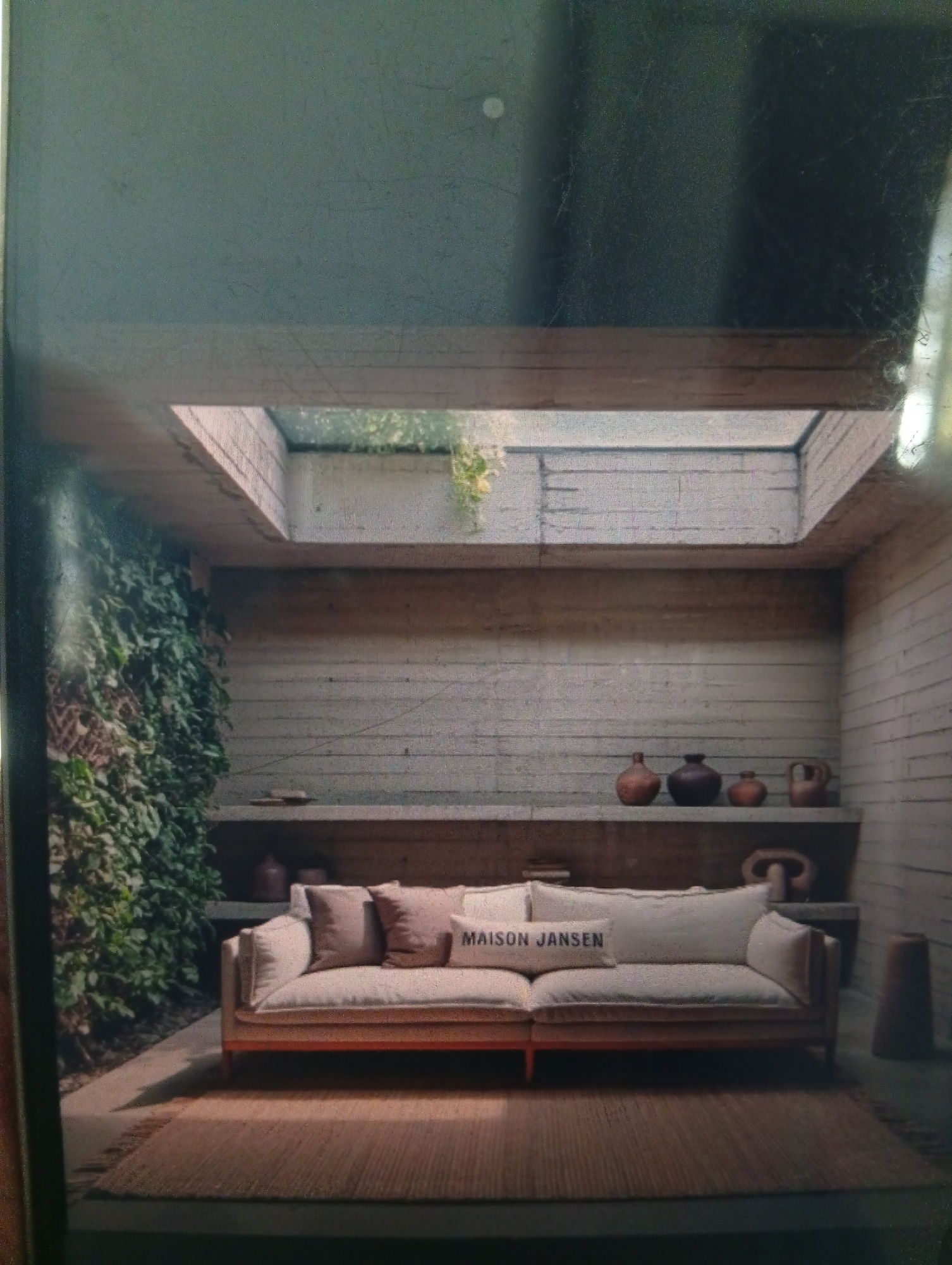 The image depicts an indoor seating area. A light-colored sofa with several throw pillows, one imprinted with "MAISON JANSEN", sits atop a brown woven rug. Behind the sofa, a long, open shelf displays various ceramic pots and other decorative objects. The walls are constructed from light-colored, possibly concrete blocks. A skylight is visible in the ceiling, allowing natural light to illuminate the space. On the left, a wall of greenery adds a natural element to the scene.
There is no information to suggest that the location is Dutsen Alhaji, Nigeria.