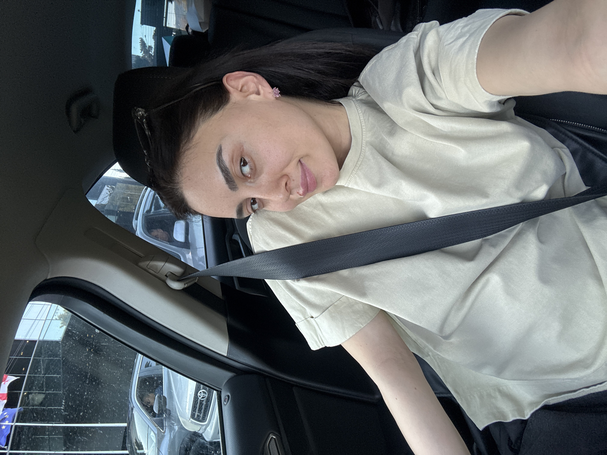 A selfie-style portrait captures a young woman seated inside a car, likely in Tbilisi, Georgia. The image is rotated clockwise, showing her head towards the left side of the frame and her body extending to the right. She has dark hair, styled back from her face, with a pair of sunglasses or eyeglasses resting on her head. Her expression is relaxed, with a slight smirk as she looks towards the camera. She is wearing a light beige or light olive green short-sleeved t-shirt and small, pink, flower-shaped earrings in her left ear. A black seatbelt is fastened across her chest and right shoulder.
The car's interior is dark, with black seats and trim visible. Through the car window, reflections of modern buildings, possibly with glass exteriors, suggest an urban environment. Another vehicle, a light-colored SUV or truck with a visible grille (resembling a Toyota emblem), is reflected clearly outside. A segment of a flag, featuring blue, white, and red colors, is also discernible outside the window. The natural light indicates the photo was taken during the daytime. The car window appears to have some dust or dirt on it. No visible text is legible within the image.