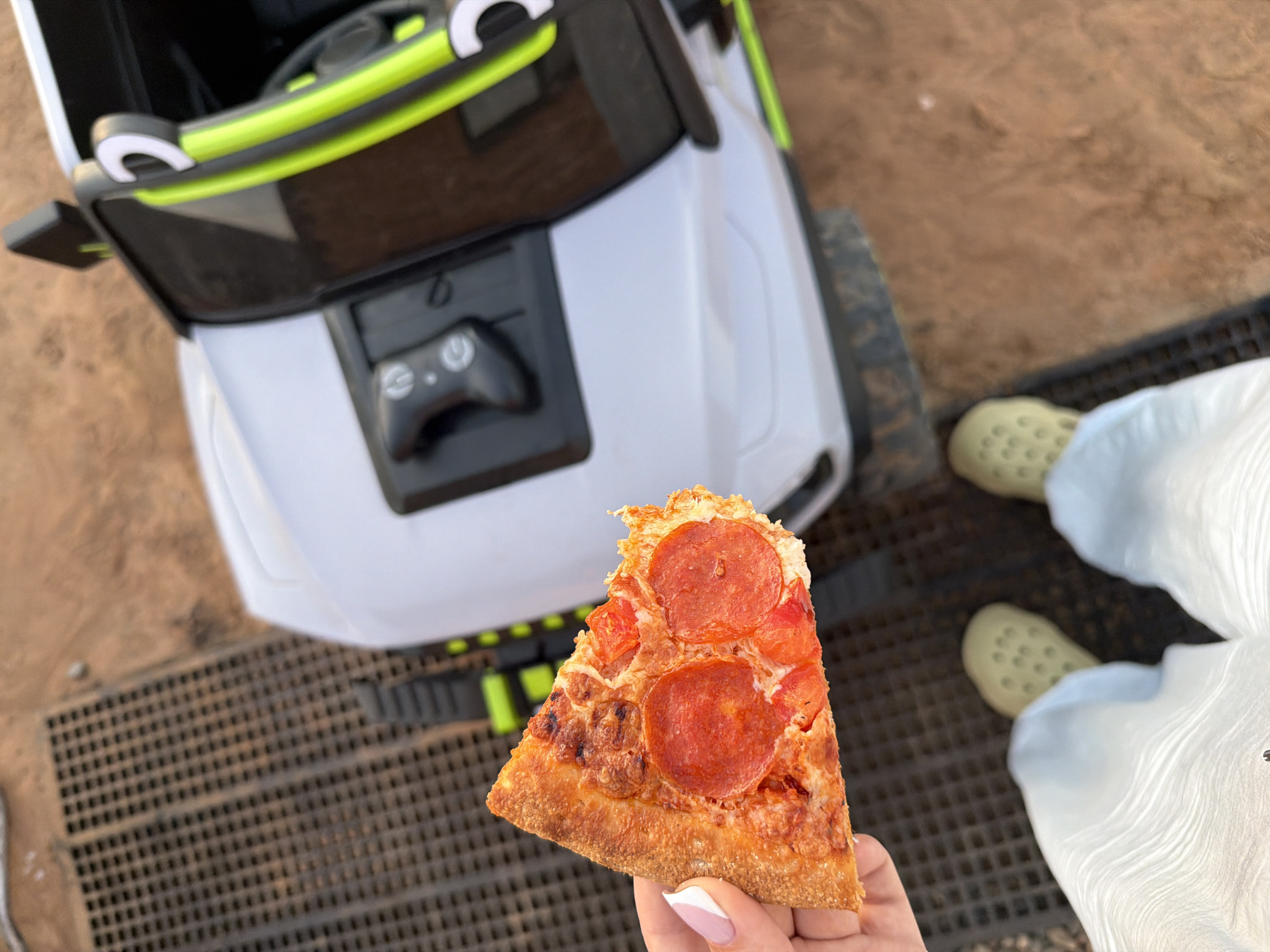 A partially eaten slice of pepperoni pizza is held in a person's hand with a pink and white manicure. The background includes a white and green child's ride-on vehicle, a metal grate, the person's feet wearing yellow clogs and white disposable booties, and a dirt surface. The exact location is impossible to determine without more information. It cannot be confirmed to be in Chaikovsky, Russia.