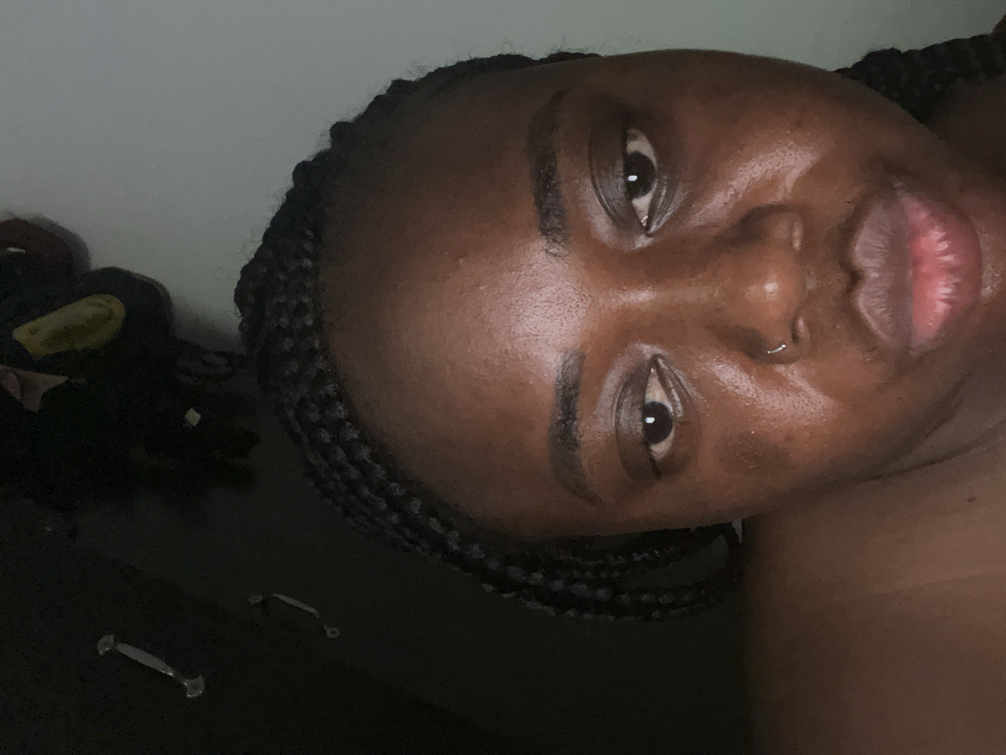 A close-up, slightly angled selfie of a dark-skinned Black woman with braided hair and a nose ring. The background includes a light-colored wall and a dark-colored surface with drawer pulls. There's no indication of Abuja, Nigeria. The woman's expression is neutral, and the lighting appears indoor.