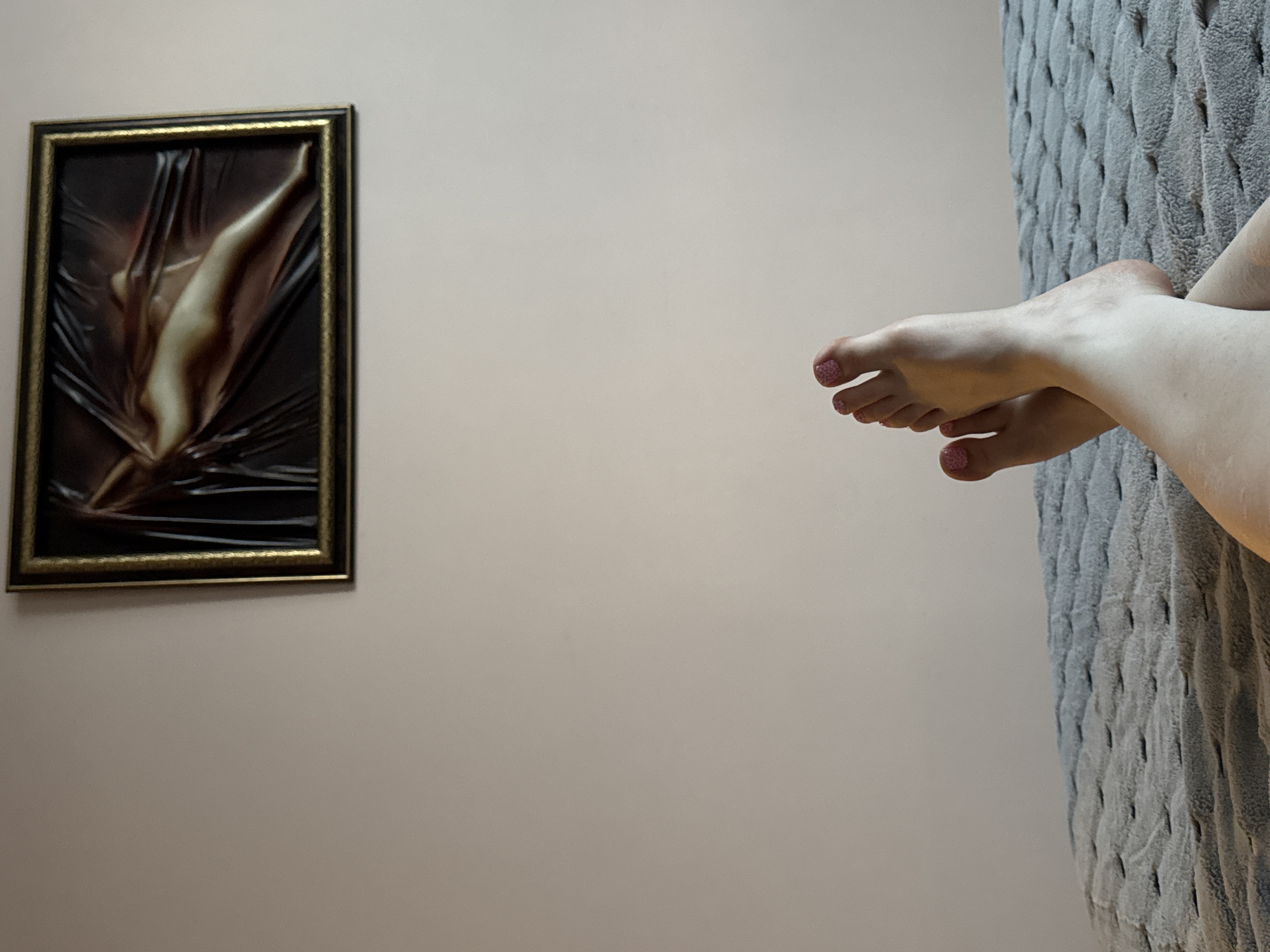 A medium-angle shot depicts a light-tan colored wall on which a dark framed painting hangs to the left. The painting displays an abstract nude figure in dark tones. To the right are a pair of pale feet with pink patterned toenail polish, presumably belonging to someone reclining on a textured gray headboard, partially visible on the right side of the image. The scene's location is unknown; there is no contextual information to suggest it is in Sochi, Russia.