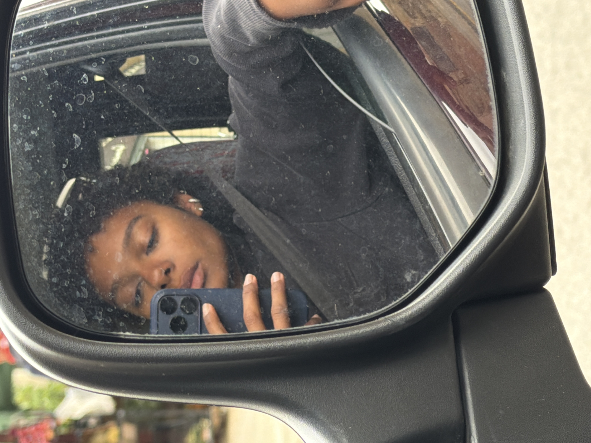 A woman is reflected in a car's side mirror. She is holding a blue smartphone, presumably taking the photograph. She is wearing a dark-colored hooded sweatshirt and has curly hair. The mirror is dirty, with water spots obscuring the view. The background outside the mirror shows a blurred, indistinct area with what appears to be green vegetation and muted colors, typical of an urban environment. The location in the city of Pals, Spain, cannot be confirmed from the image.