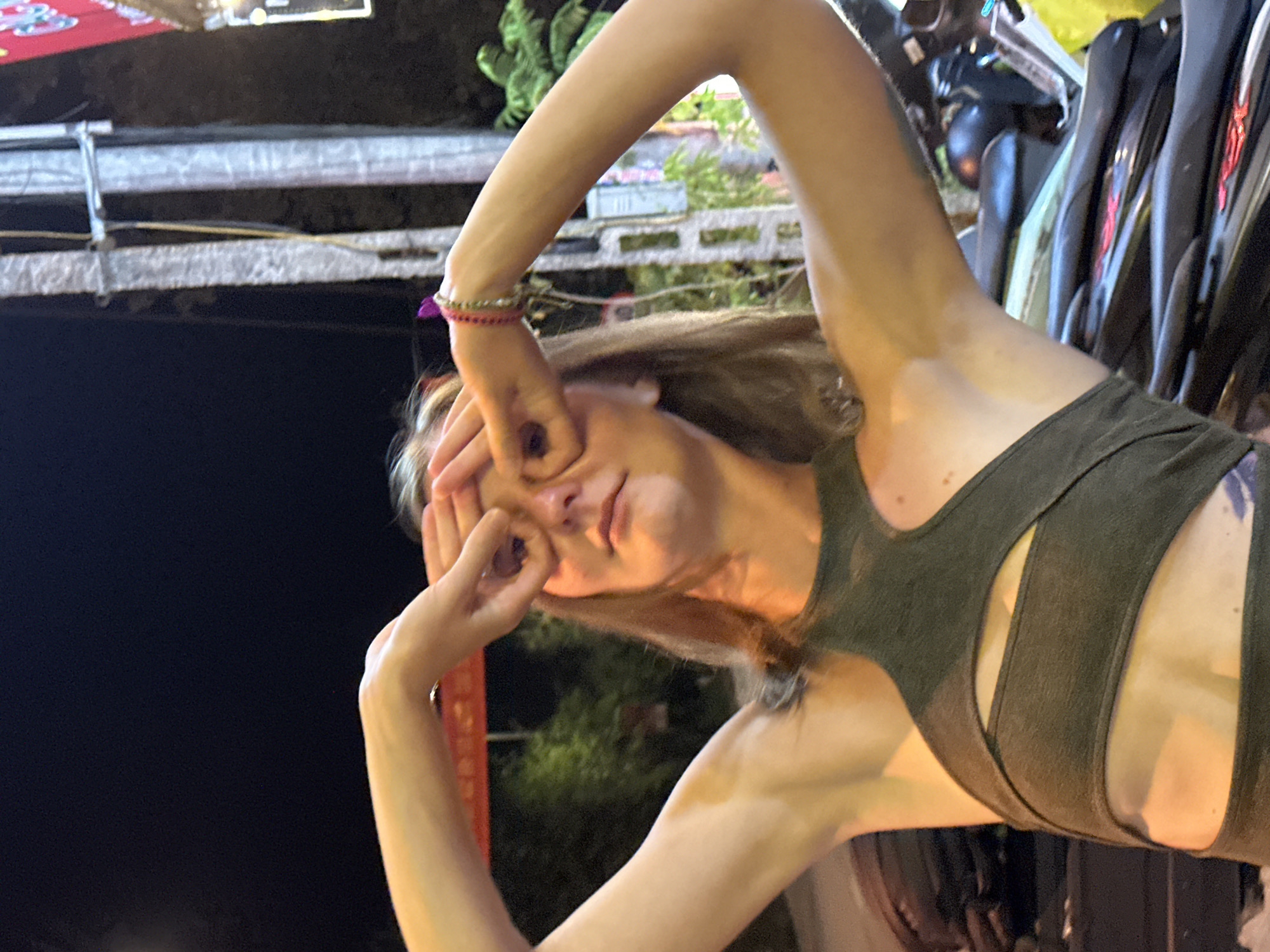 The image features a young woman posing outdoors at night in Mù Cang Chải, Vietnam. She has light brown hair and is wearing a dark green or olive-colored sleeveless top with a unique strap design. On her right wrist, she wears a multi-strand bracelet featuring pink, red, and silver beads. Her arms are raised, and her hands are positioned to create circles around her eyes, as if looking through imaginary binoculars or spectacles, with her gaze directed towards the camera. Her head is tilted, and her expression appears neutral to slightly playful.
The scene is illuminated by warm, artificial lighting, casting a yellow-orange glow on her skin and creating soft shadows. The background is dark, indicating the nighttime setting. Blurred artificial lights are visible in the distance, along with indistinct green foliage. To the upper left, a light-colored horizontal beam or structure is present. On the right side of the frame, several dark, glossy objects resembling the seats or bodywork of motorcycles are stacked vertically, suggesting a parking area or a busy street setting. A partial reddish banner or sign is vaguely discernible in the left background, but no text is clearly visible on any object.