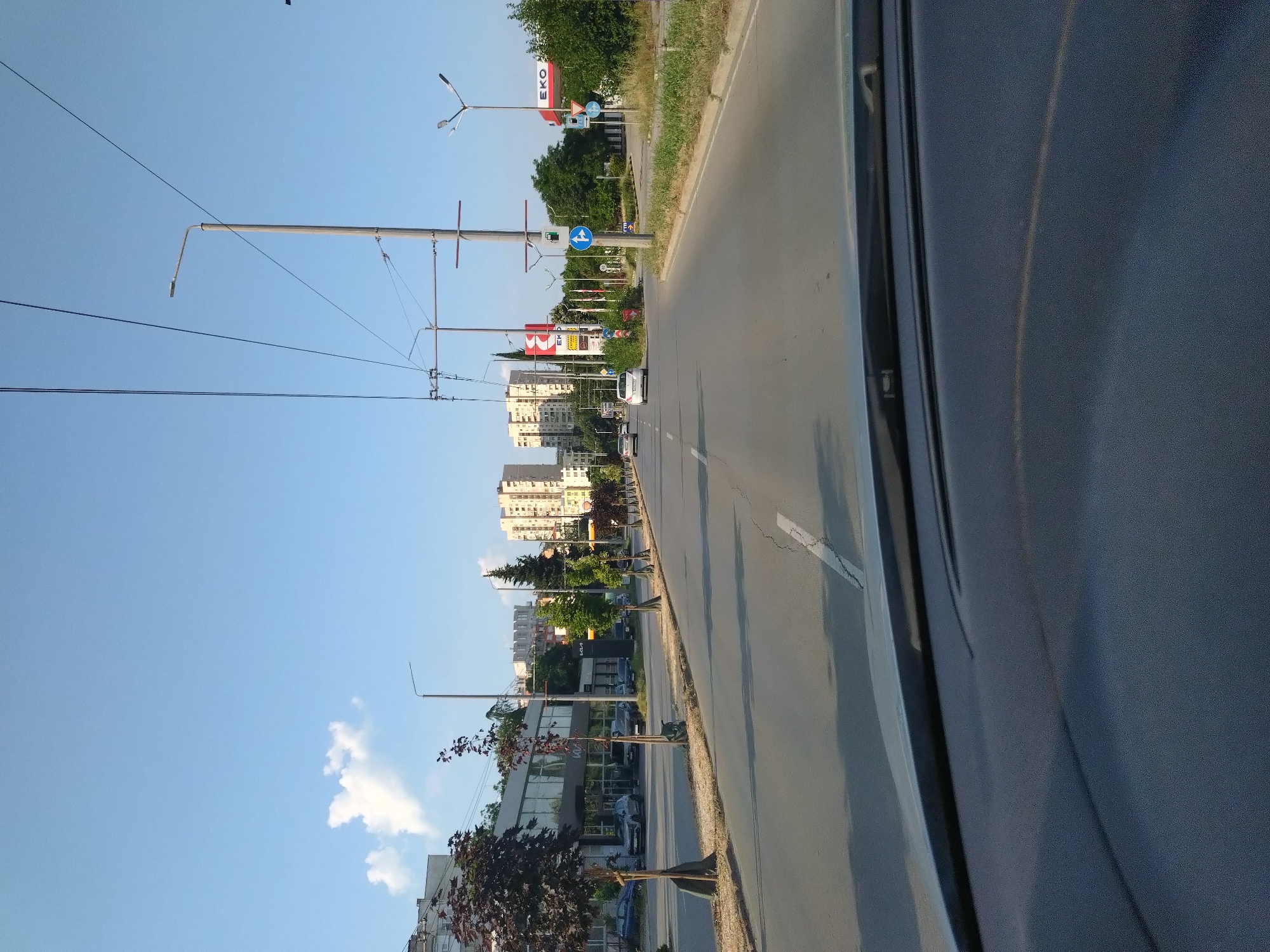 The media file captures a street scene in Stara Zagora, Bulgaria, likely during the daytime. The image is oriented vertically, showing a road with passing cars and a sidewalk lined with trees and buildings. Streetlights and overhead wires are prominent features. On the right side of the frame, a dark, textured surface, possibly the interior of a vehicle, partially obstructs the view.
The buildings visible are modern, multi-story structures, some appearing to be apartment blocks and others commercial establishments, including a "KIA" dealership. There are also signs for "EKO," suggesting a gas station. Traffic lights and road signs are present, including directional signs. The sky is clear and blue with a few scattered clouds. The overall impression is that of a typical urban environment on a sunny day. No people are clearly discernible in the image. The scene suggests normal daily activity, with vehicles moving along the road. The lighting indicates it's not early morning or late evening.