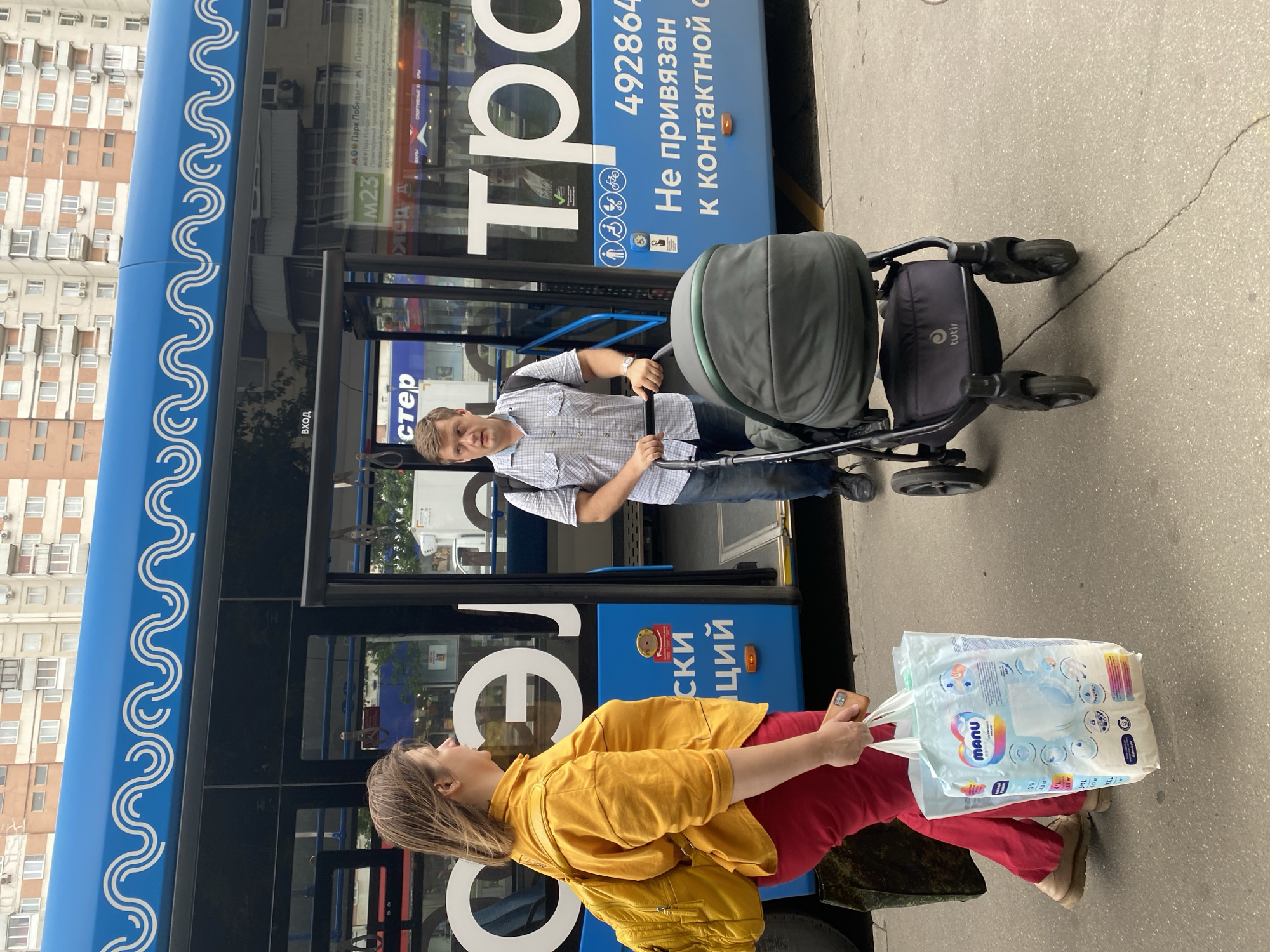 A woman wearing a yellow shirt and red pants is standing on a sidewalk next to a blue bus, holding a plastic bag filled with diapers. A man in a checkered shirt is standing behind her, pushing a stroller. The bus has text on its side that appears to be in Russian. The background shows apartment buildings. The scene is likely in Moscow, Russia, given the Cyrillic text on the bus and the style of the buildings.