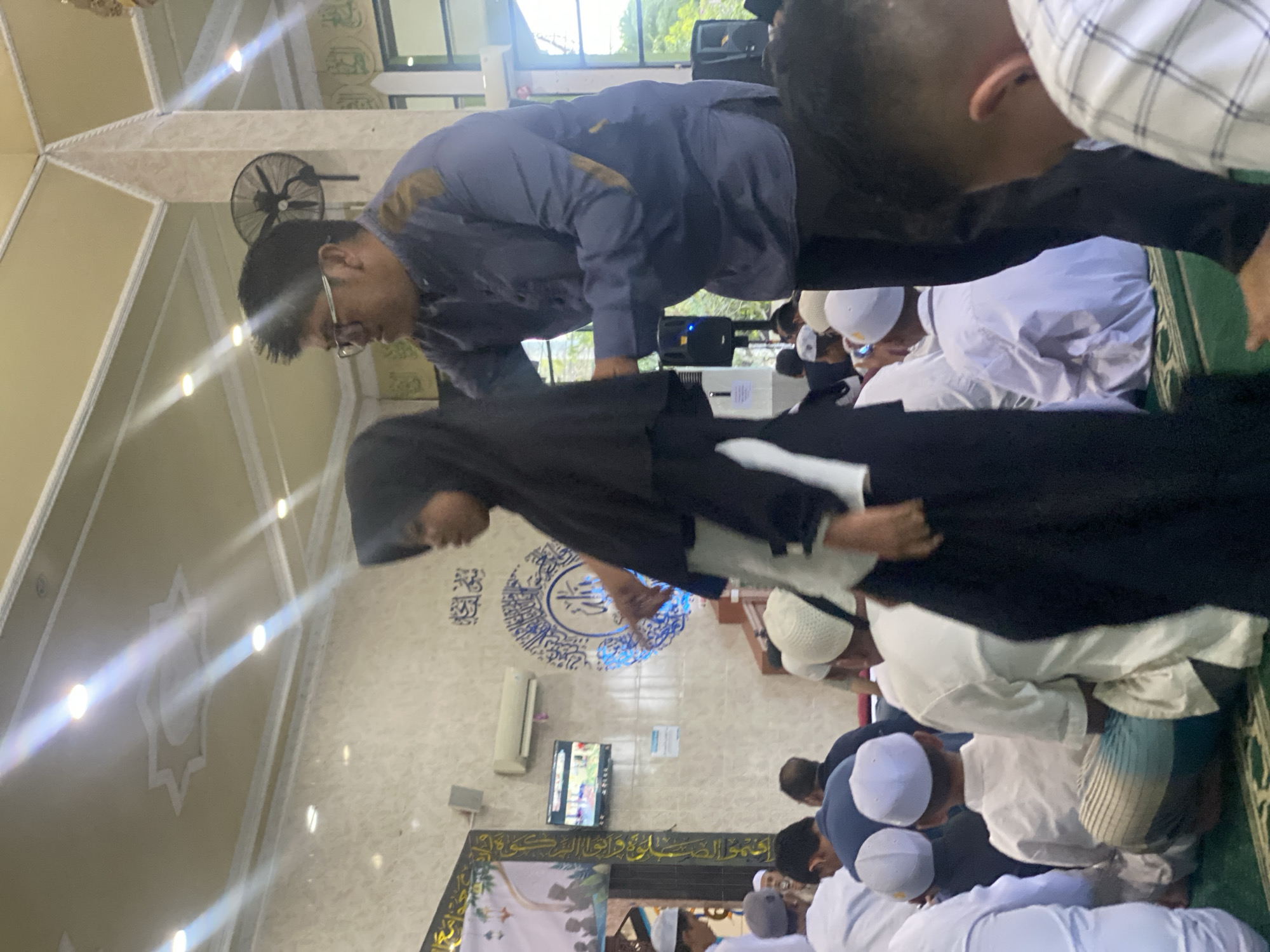 The image shows an indoor gathering, likely a religious event, taking place in the city of Banjarbaru, Indonesia. The environment appears to be a prayer hall or mosque, indicated by the presence of prayer mats on the floor, religious calligraphy on the walls, and what appears to be a mihrab or prayer niche in the background. The ceiling features decorative star patterns and spotlights. A prominent large pillar, possibly structural or decorative, stands near the center.
In the foreground, a young female wearing a black hijab and a long dark dress is partially visible on the left, facing right. To her right, an adult male wearing glasses, a blue shirt with gold accents on the sleeves and collar, and dark pants is seen from the side, reaching towards the girl. Several other individuals, predominantly males of varying ages, are seated on the floor, facing towards the front of the room. Many are wearing white prayer caps and traditional Islamic attire.
On the wall in the mid-ground, a television screen is mounted, displaying colorful imagery. Above it, an air conditioning unit is visible. A black oscillating fan is mounted on the wall behind the male figure. The overall scene suggests a communal gathering with an atmosphere of devotion and order.