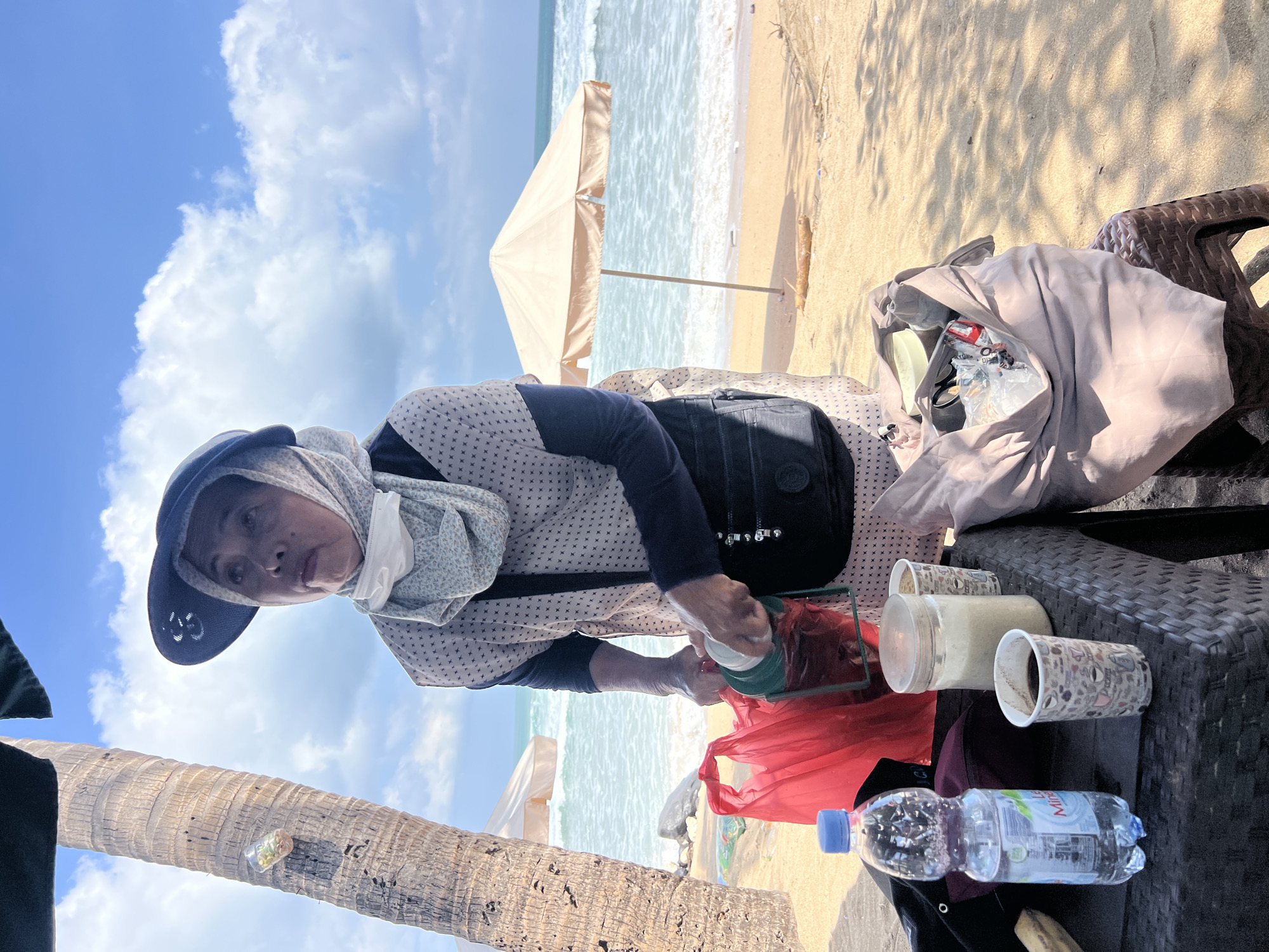 A middle-aged woman wearing a blue wide-brimmed hat and a patterned hijab is standing on a sandy beach, likely in Kuta, Indonesia. She is looking upwards and to her left, with a surgical mask pulled down below her nose. In front of her, on a woven plastic table, are several items: a bottle of "Le Minerale" water, two paper cups with beverages (one dark, likely coffee, and one lighter, possibly with sugar or creamer), and a clear jar filled with white granular material. The woman is holding a green and white insulated container and appears to be preparing or serving something. To her left, a bright red plastic bag is visible. Behind her, the ocean waves are rolling onto the shore, and a light-colored beach umbrella stands further down the beach. A palm tree trunk is to her left, and a brown plastic stool with a tan cloth bag filled with various items is to her right. The sky is blue with scattered white clouds.