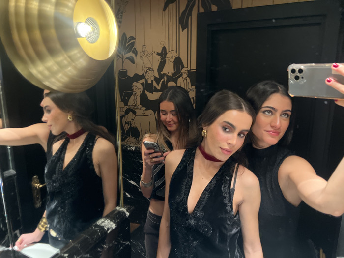 Three women pose in a bathroom. Two are in the foreground, the woman on the left wears a velvet choker and a sleeveless top with decorative embellishments. She is partially reflected in a mirror. The woman on the right holds up a phone, presumably to take a selfie. Behind them and slightly out of focus is a third woman looking at her own phone. The bathroom features a large brass light fixture, dark walls, and a decorative mural in the background.