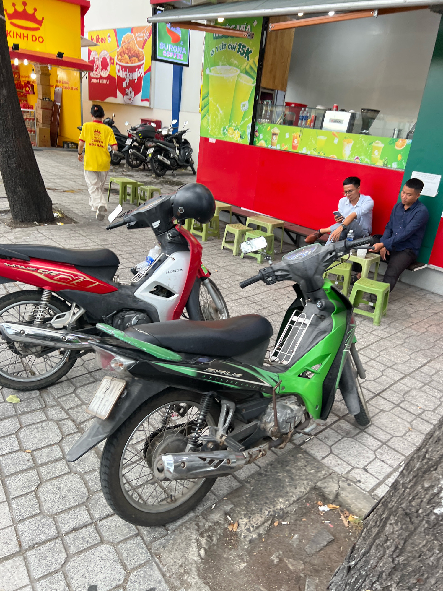 In Nha Trang, Vietnam, on a pedestrian walkway lined with trees and adjacent to a street, two men are seated on plastic stools outside a food stall. One man, wearing glasses and a light blue collared shirt, is looking down at his phone. The other man, in a dark blue collared shirt, sits with his arms crossed. Two motorcycles are parked on the walkway; one is red and silver, and the other is green and black. In the background, a food stall with a red and green facade displays advertisements for "Jollibee" and "SURONA COFFEE." A person in a bright yellow shirt walks away from the stall, carrying what appears to be a delivery bag. The ground is paved with gray interlocking bricks, and some litter is visible near the base of a tree.