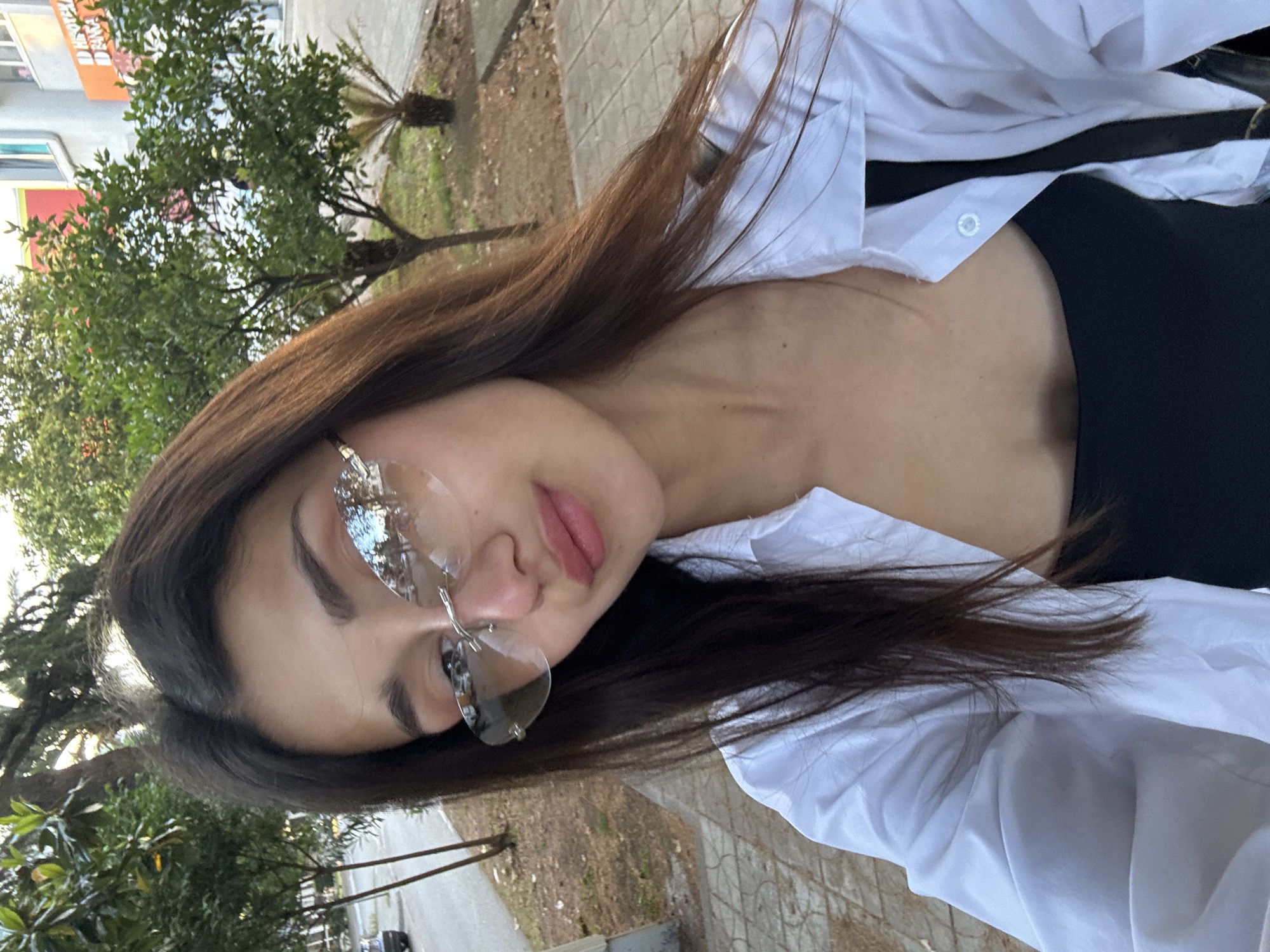 A young woman with long brown hair, wearing rimless glasses, a black top, and a white button-down shirt, is seen from a high angle. She appears to be on a sidewalk or paved area, with trees and a building visible in the background. The scene is likely located in an urban setting, specifically in Bar, Montenegro, based on the context provided. The woman is the primary subject, and the environment includes typical city elements like sidewalks, buildings, and trees.